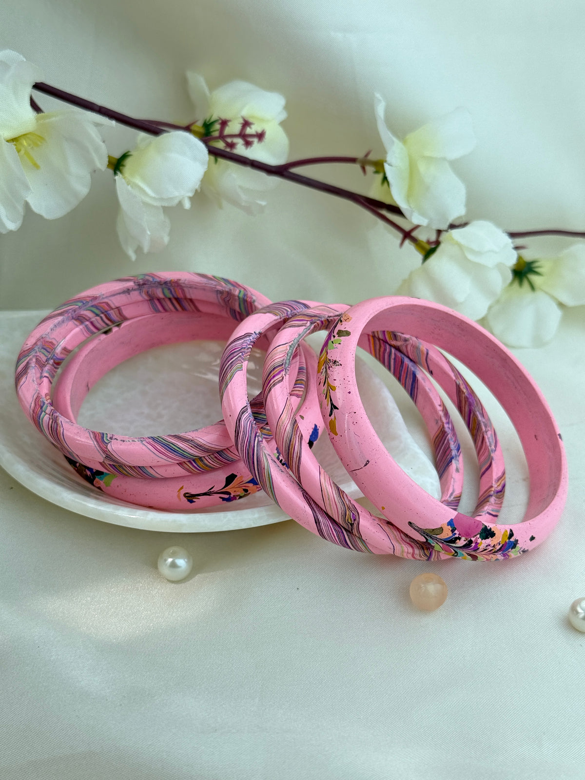 A set of pink bangles, including solid pink and patterned pink-and-purple bangles, arranged on a white plate with decorative white flowers in the background.