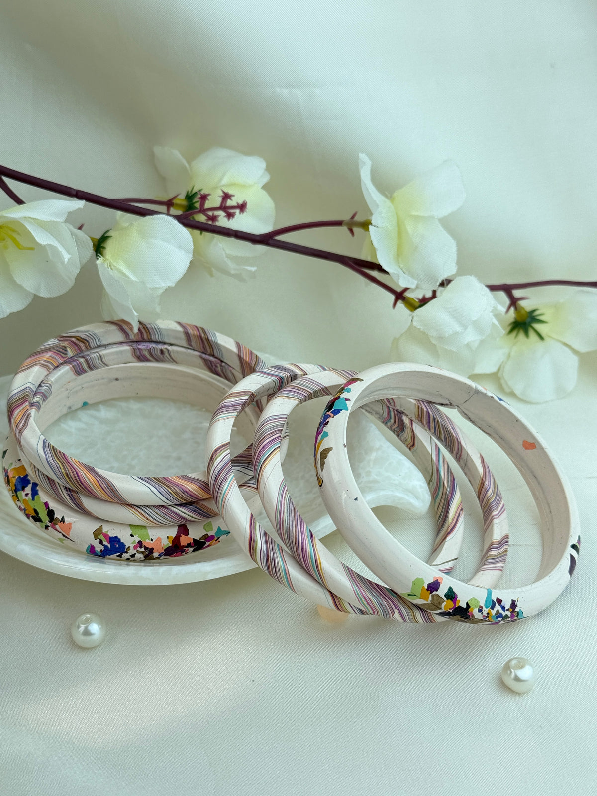 A set of handmade bangles with a cream base and colorful floral/striped patterns, arranged on a white plate with decorative white flowers in the background.