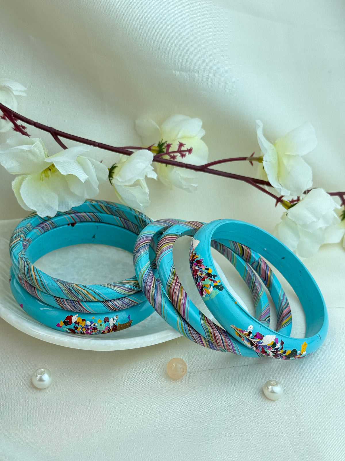 A set of turquoise blue bangles with colorful striped and floral patterns, displayed on a white plate with decorative white flowers and small pearls.