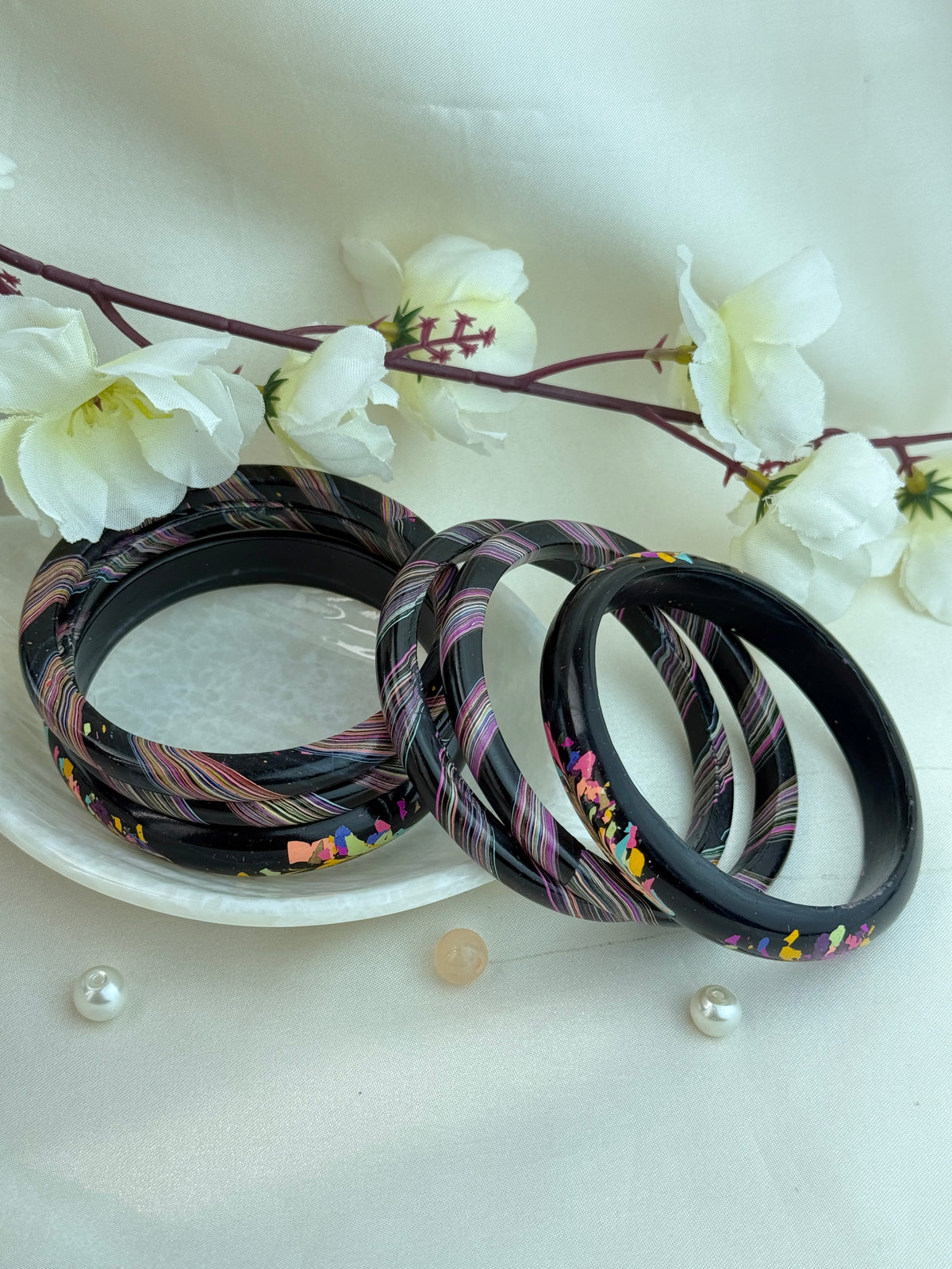 A set of black bangles with colorful striped and speckled patterns, arranged on a white plate with white flowers and small pearls in the background.