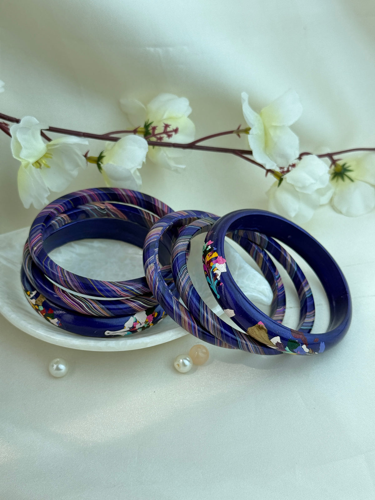 A set of dark blue bangles with colorful striped and floral patterns, displayed on a small white dish with decorative white flowers in the background.