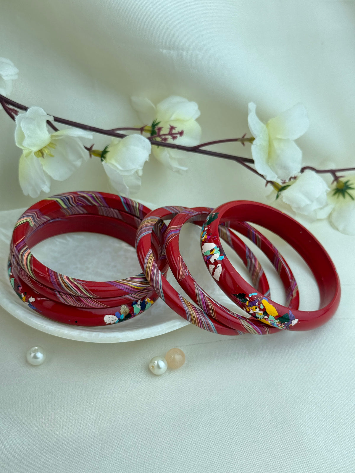 A set of red bangles with colorful striped and floral inlay patterns, arranged on a white plate with decorative white flowers and small pearls against a soft white background.