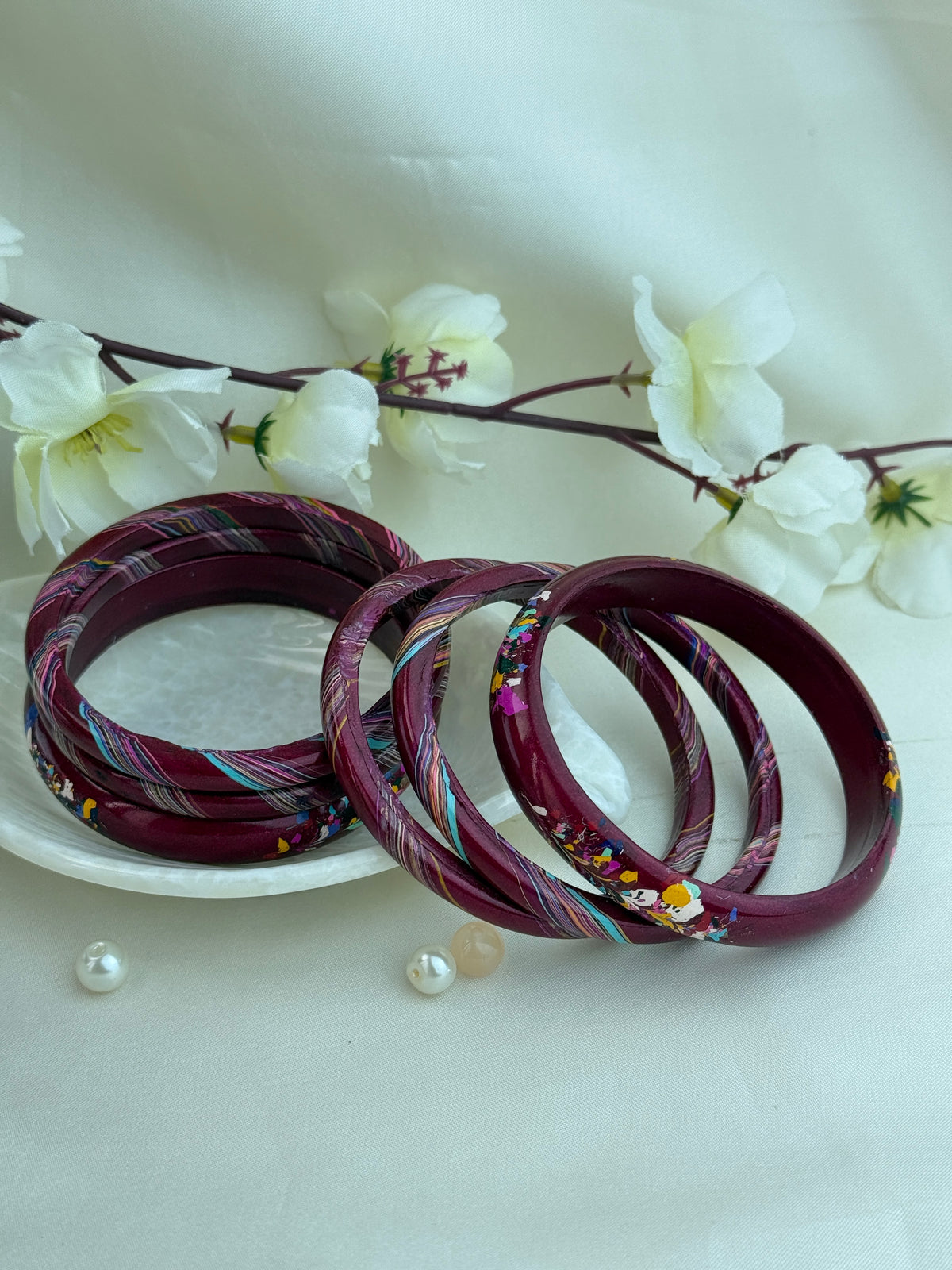 Four maroon bangles with colorful woven patterns and small floral details, arranged on a white plate with white flowers in the background.