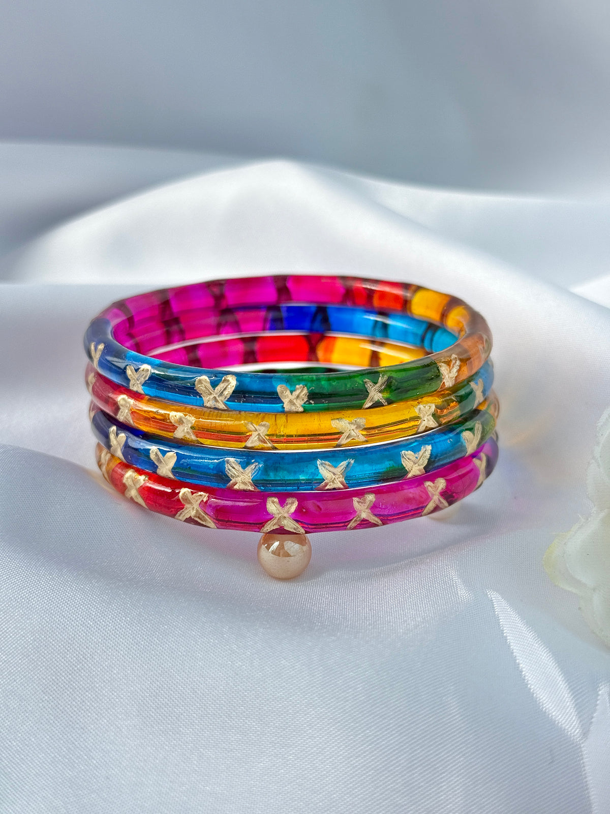 A stack of four colorful translucent bangles with gold butterfly details and a small gold bead, displayed on a white satin fabric background.