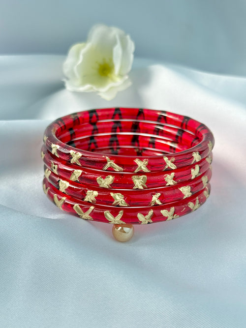 A stack of red bangles with black geometric patterns and gold bow motifs, displayed on a white satin fabric with a white flower in the background.