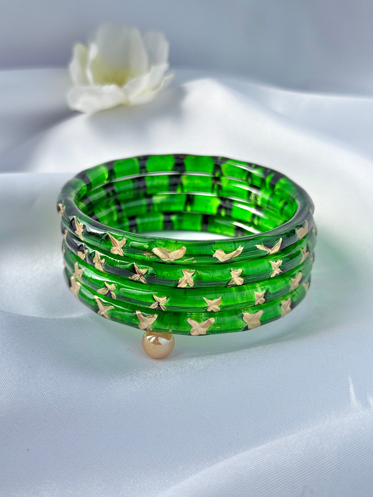 A stack of green glass bangles with black grid patterns and gold butterfly details, displayed on a white satin fabric background with a small white flower in the background.