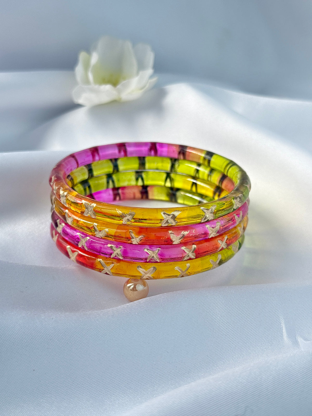 A stack of four translucent bangles in pink, orange, yellow, and green with black stripes and small gold butterfly details, displayed on a white satin fabric background with a white flower in the background.