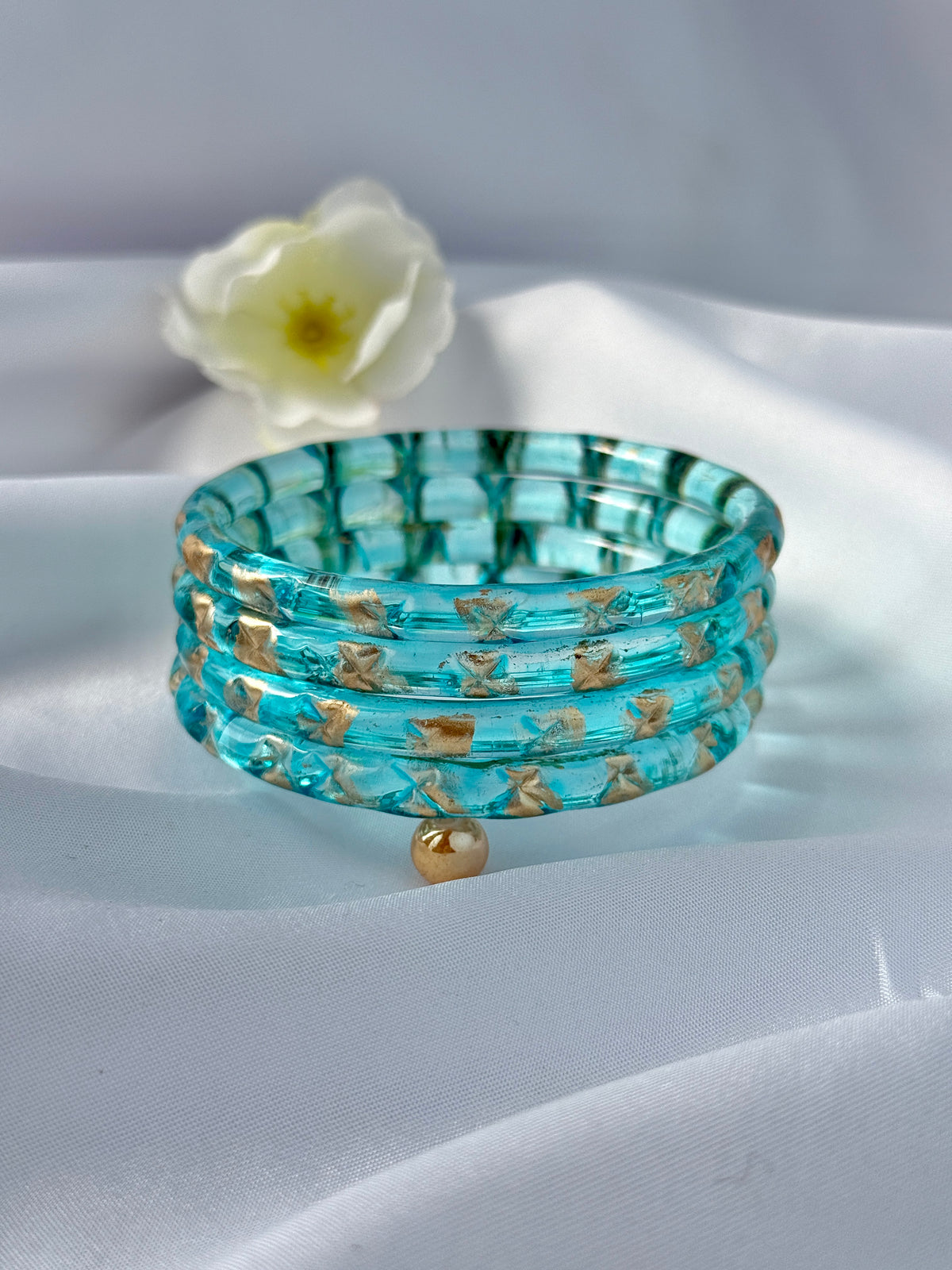A turquoise blue beaded wrap bracelet with small gold star accents, displayed on a white draped fabric background with a white flower prop.