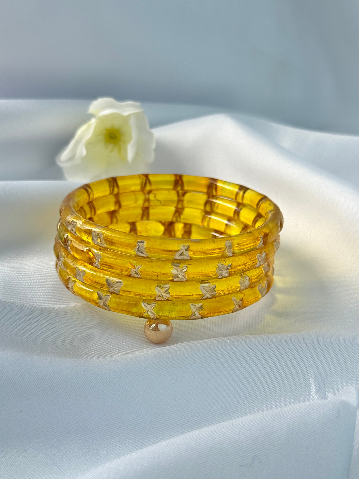 A stack of five translucent yellow bangles with a grid pattern and small silver X-shaped accents, displayed on a white satin fabric background with a white flower prop.