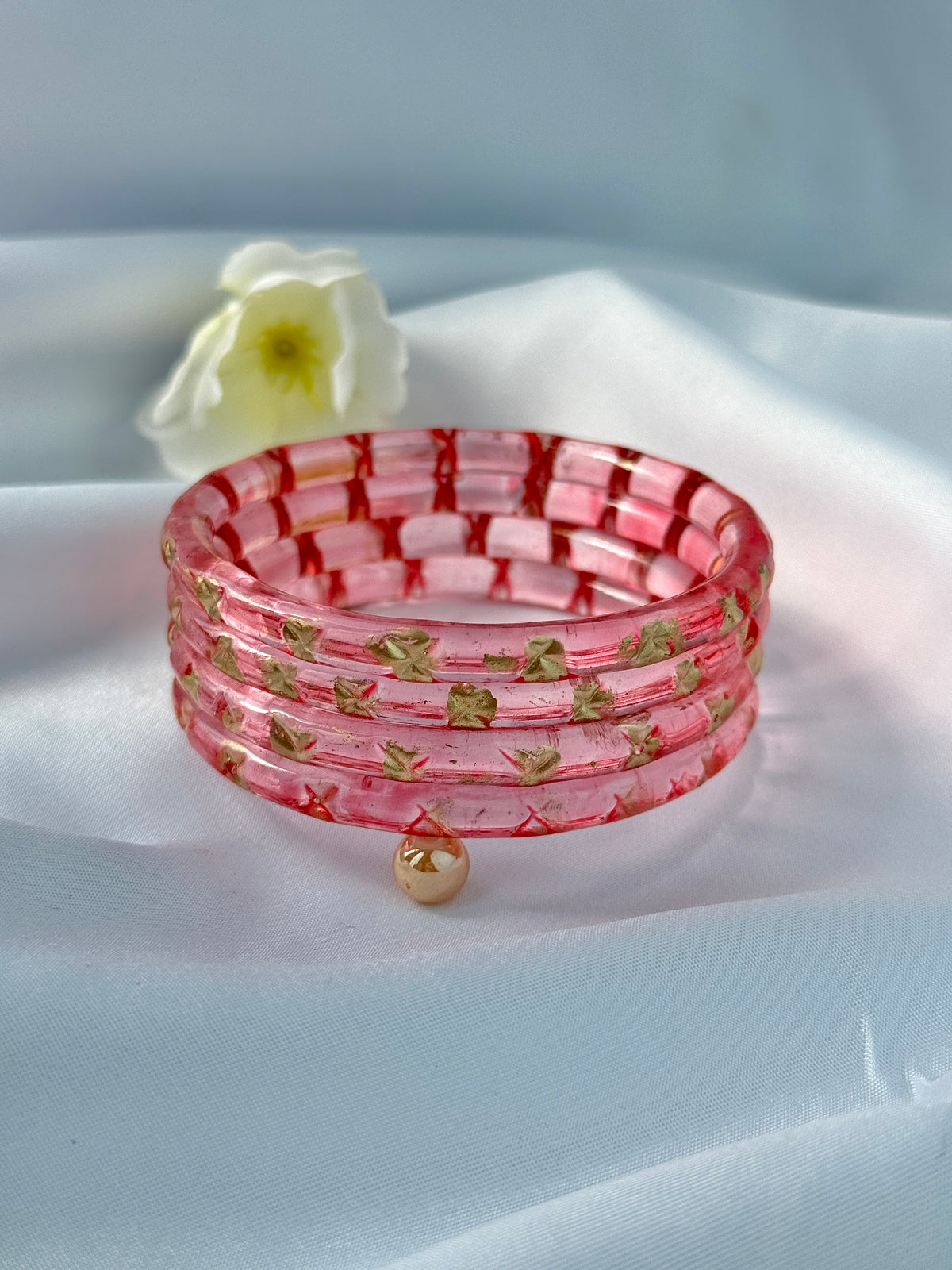 A pink translucent bangle bracelet with a grid pattern and small gold leaf details, resting on a white satin fabric background with a white flower prop.