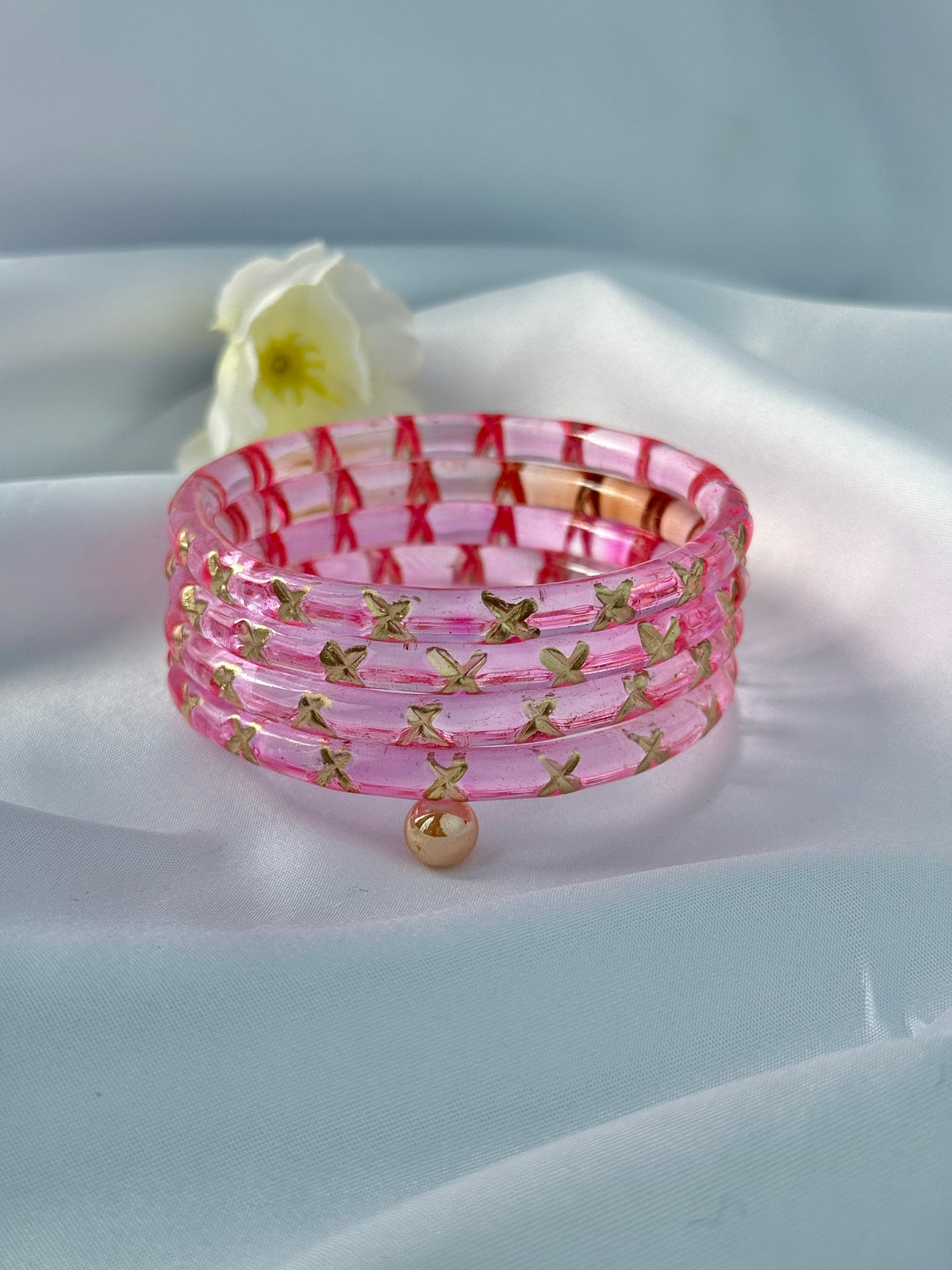 A stack of five pink translucent bangles with gold butterfly motifs and a small dangling gold bead, displayed on a soft white fabric background with a white flower prop.