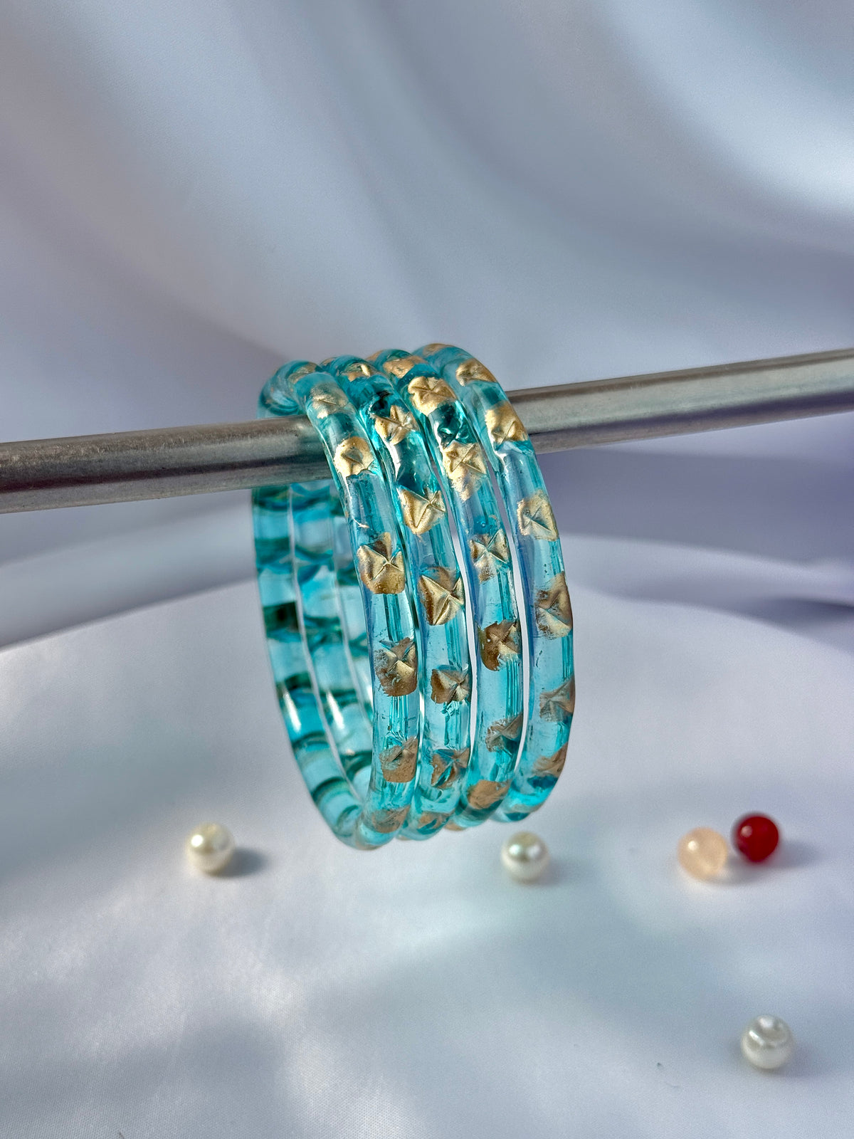 A set of five stacked turquoise blue bangles with gold foil butterfly patterns, hanging from a metal rod against a draped white fabric background.