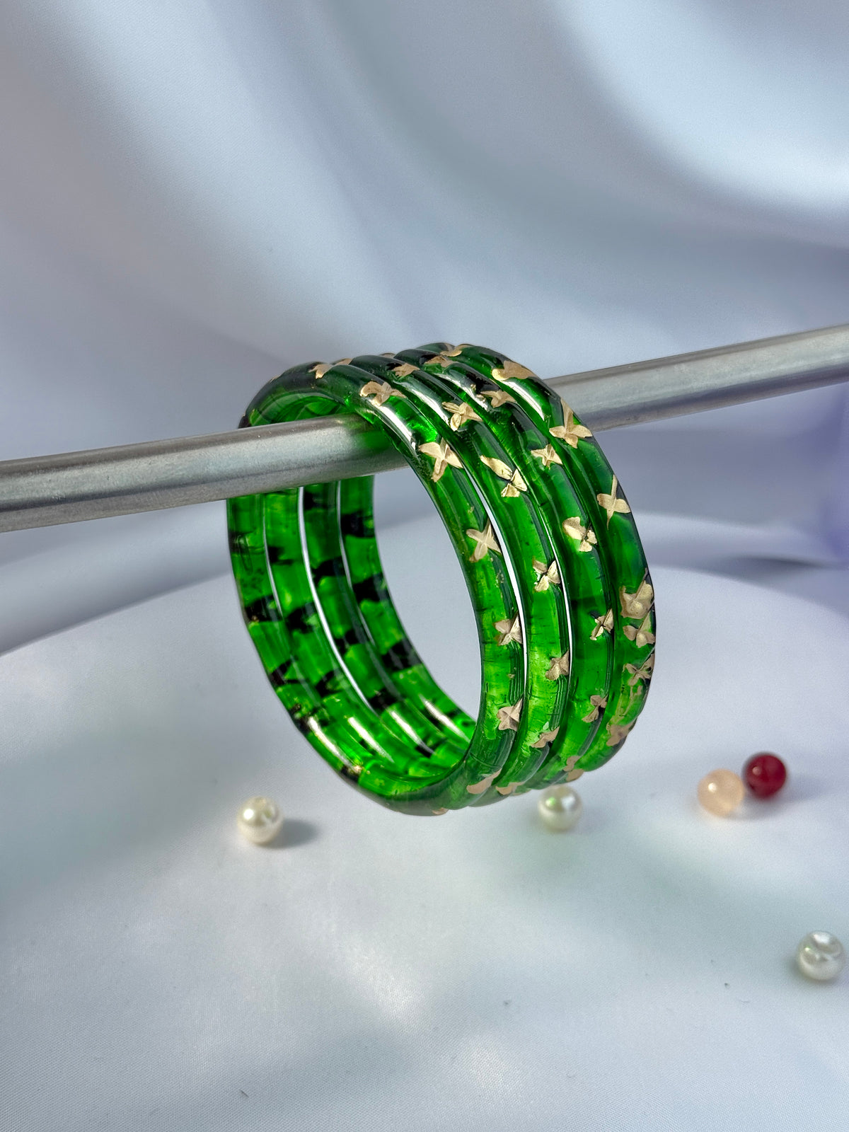 A stack of five green glass bangles with gold and black floral patterns, resting on a silver rod against a soft white fabric background.