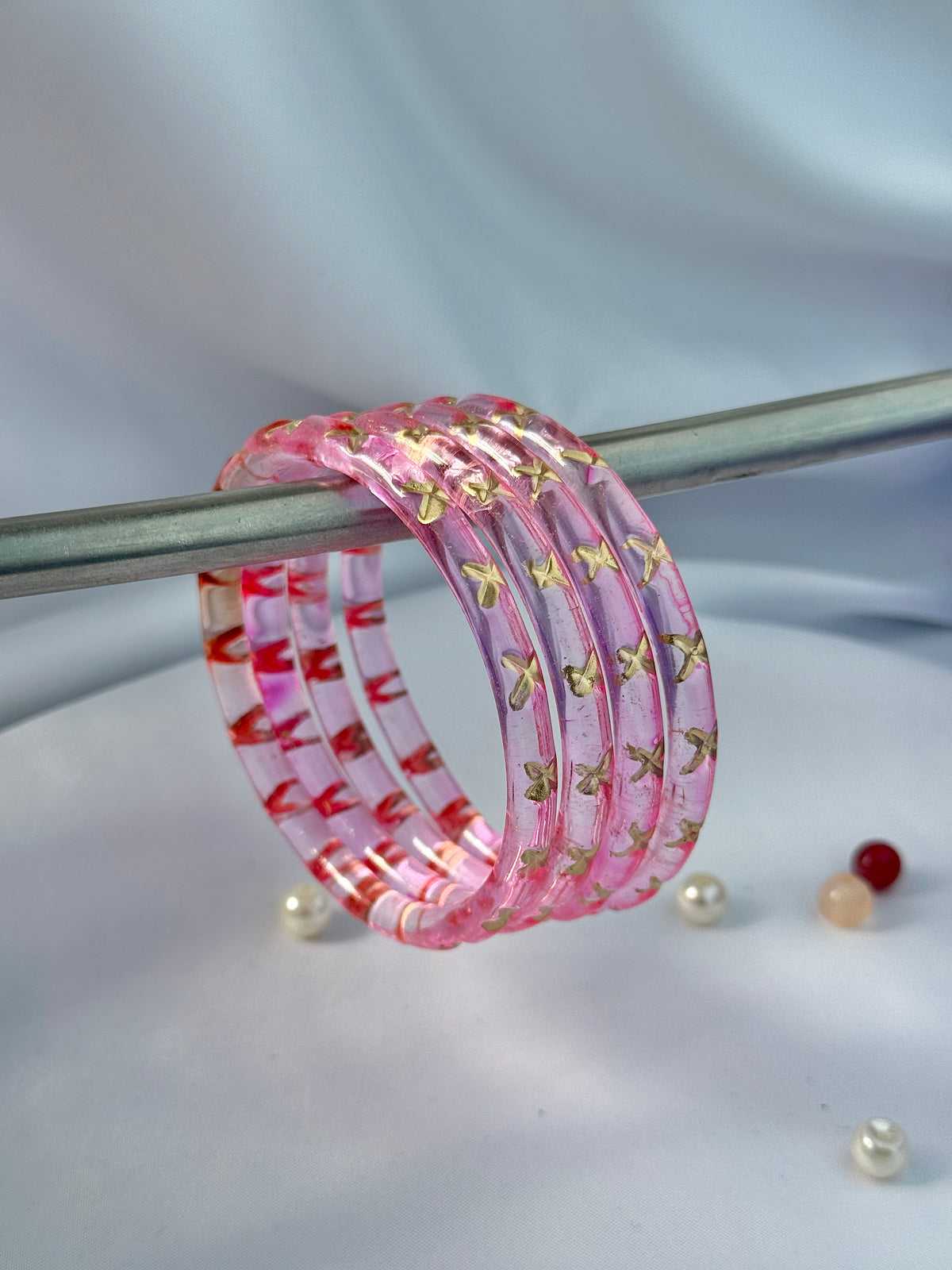 A stack of pink translucent bangles with gold butterfly motifs and red floral accents, draped over a metal rod on a light fabric background.