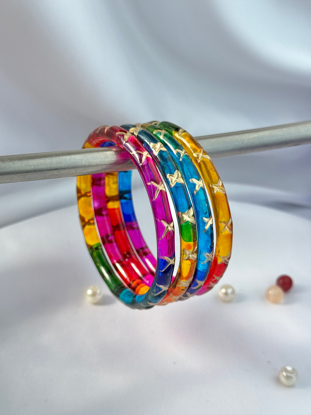 A set of five colorful bangles with gold star patterns, displayed on a silver rod against a white draped background.