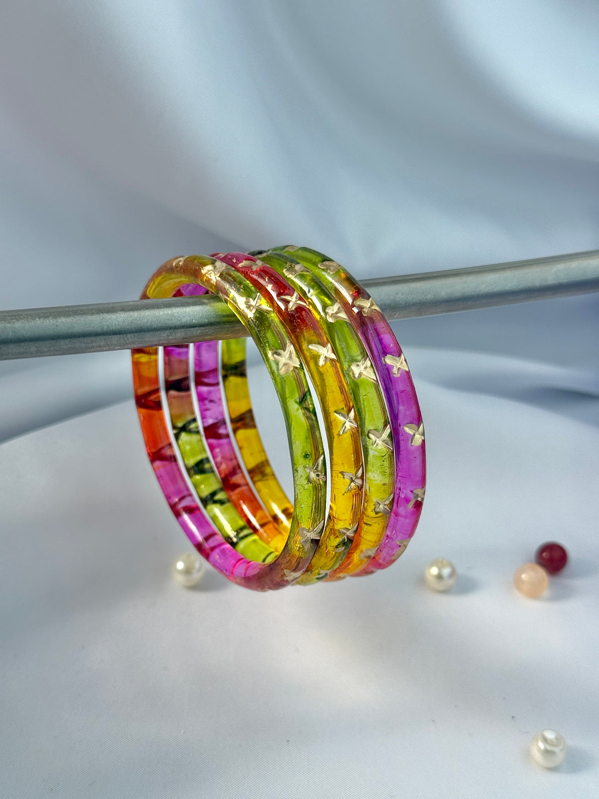 A set of five colorful bangles with gold star patterns, displayed on a metal stand against a light gray draped fabric background.