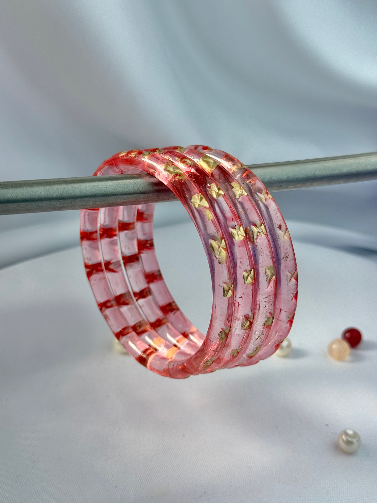 A set of five translucent pink glass bangles decorated with gold foil stars and red stripes, displayed on a metal stand.