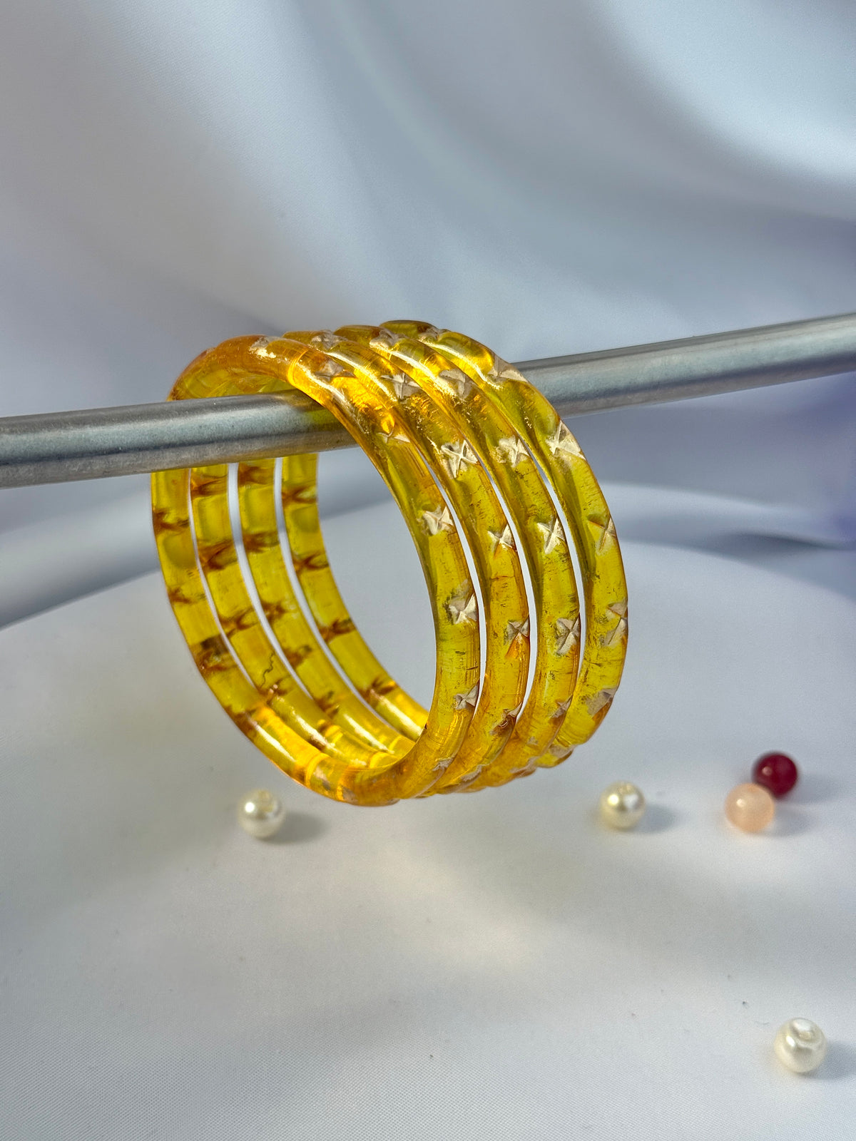 A set of five stacked translucent yellow bangles with a textured pattern and small butterfly details, displayed on a silver rod against a draped white fabric background.