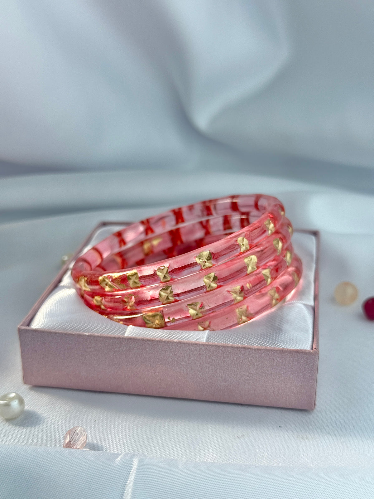 A stack of five translucent pink bangles decorated with small gold star-shaped charms, presented in a pink gift box on a soft white fabric background.
