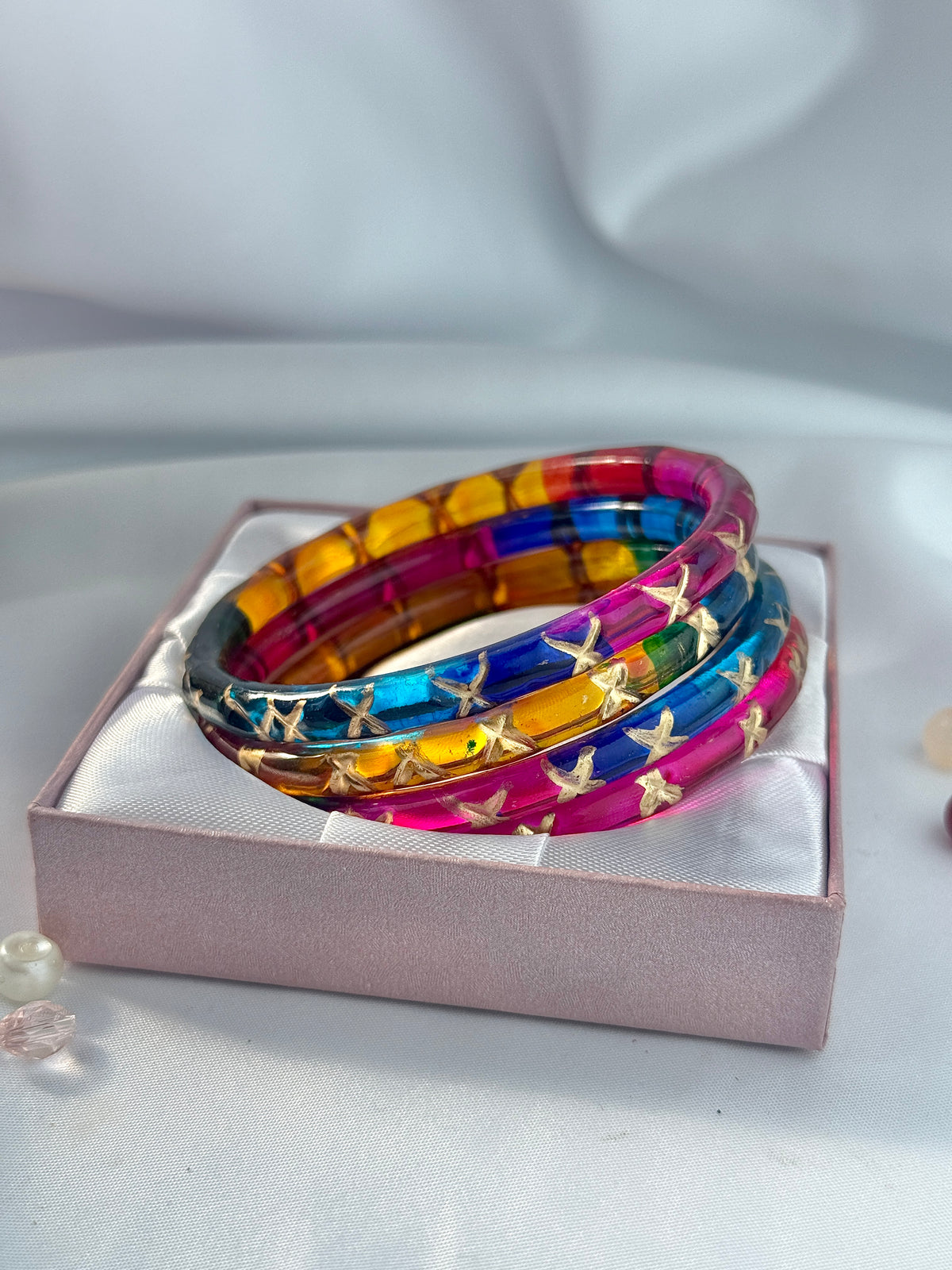 A stack of four colorful bangles with gold bird motifs, presented in a pink gift box on a soft white fabric background.