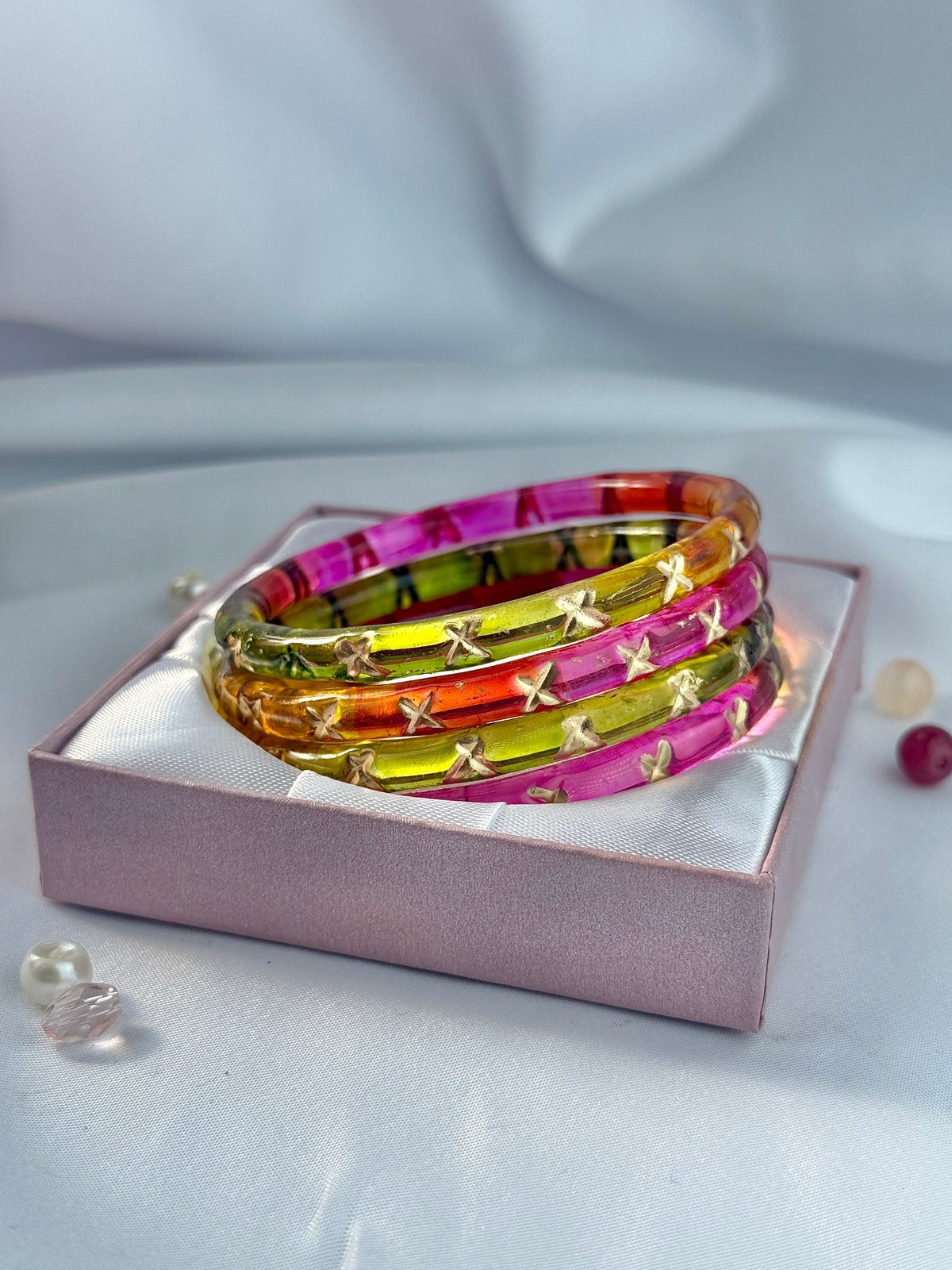 Four stacked translucent bangles in pink, orange, yellow, and green, each decorated with small gold star motifs, displayed on a pink gift box.