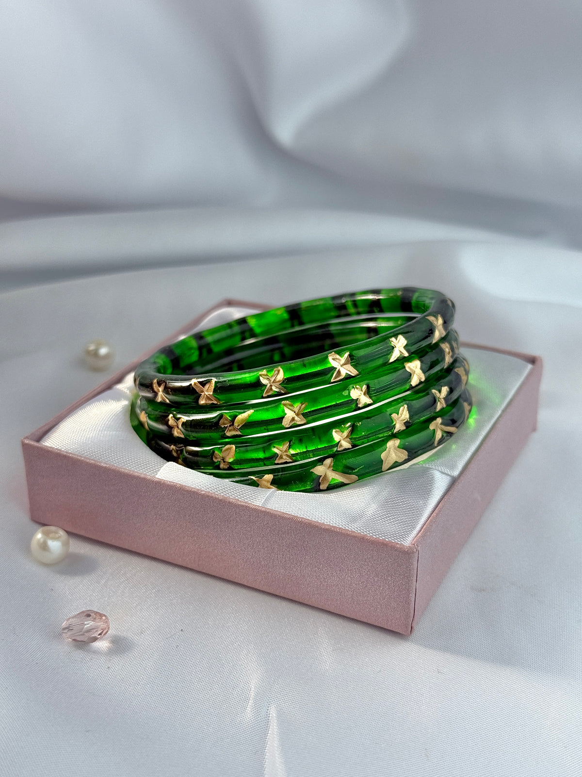 A stack of five green bangles with small gold butterfly embellishments, presented in a pink gift box on a white satin background.