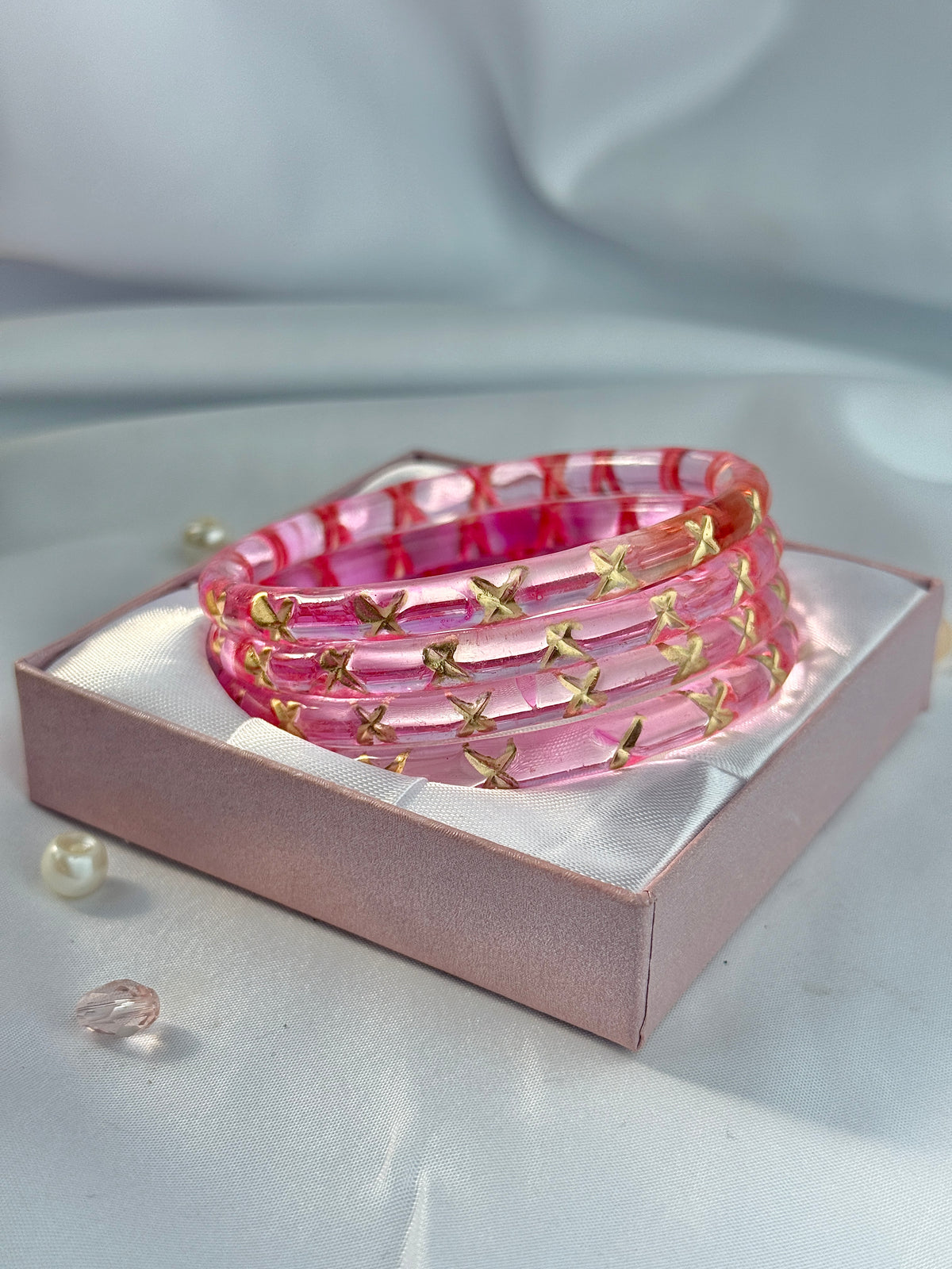 A stack of pink resin bangles with gold butterfly embellishments, presented in a pink gift box on a draped satin fabric background.