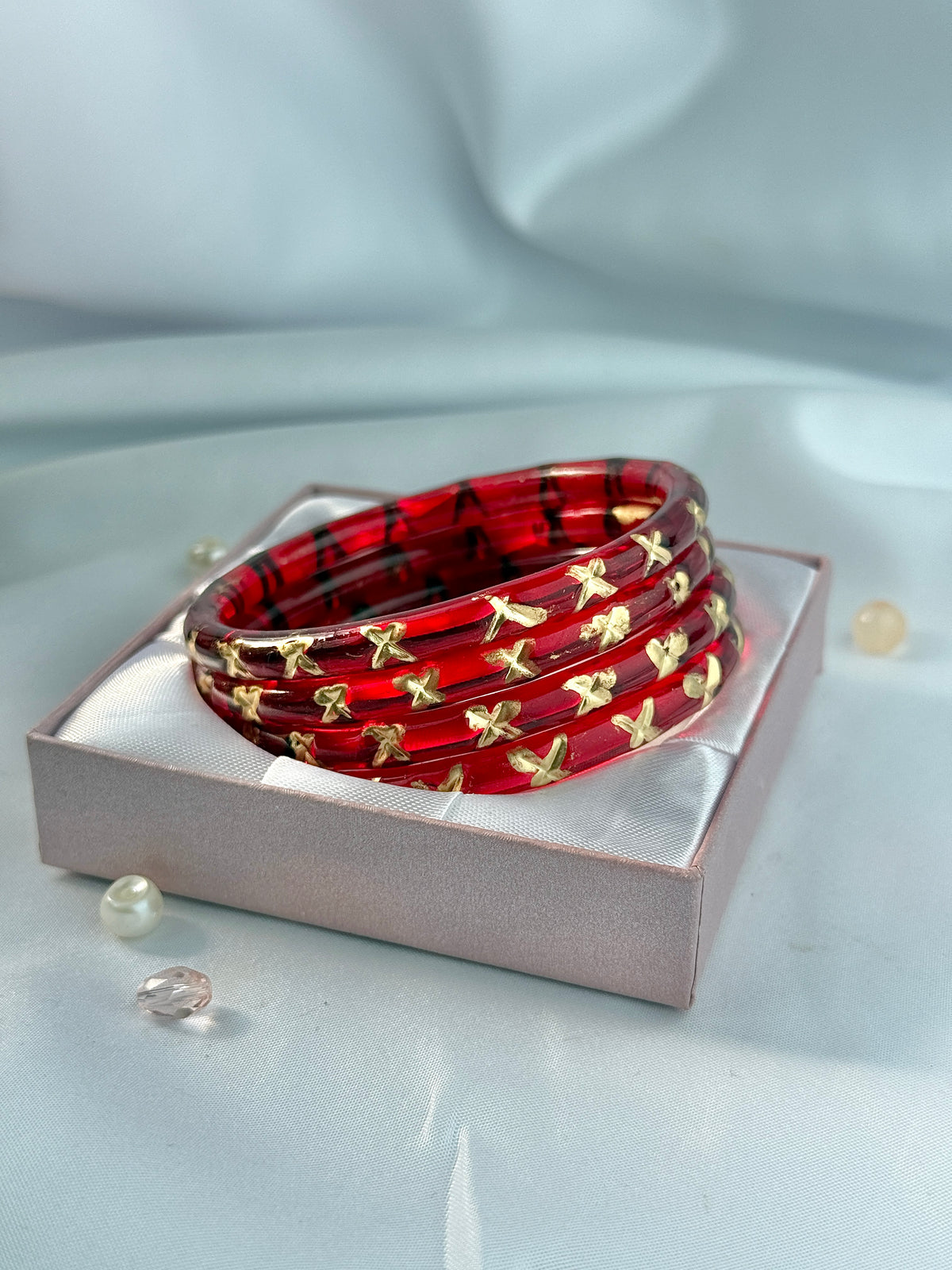 A stack of red bangles with gold floral motifs, presented in a light pink gift box on a soft, light gray draped fabric background.
