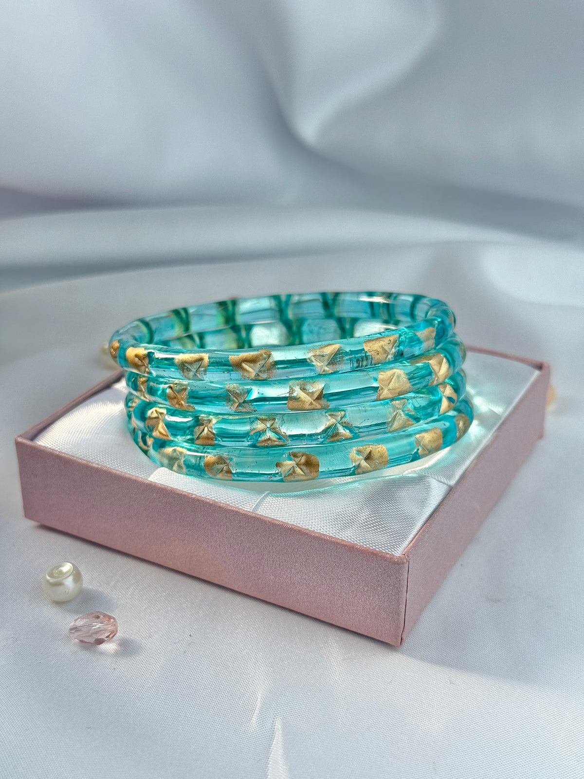 A stack of translucent aqua blue bangles with small gold decorative accents, displayed on a pink gift box with a white satin lining. The box is set against a softly draped white fabric background.