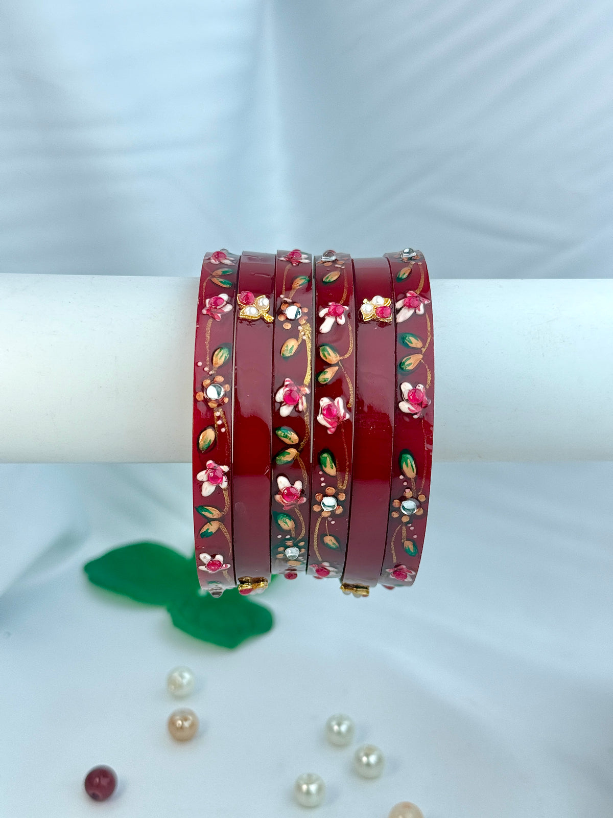 Four red bangles with floral and paisley patterns, displayed on a white cylindrical stand against a white fabric background.