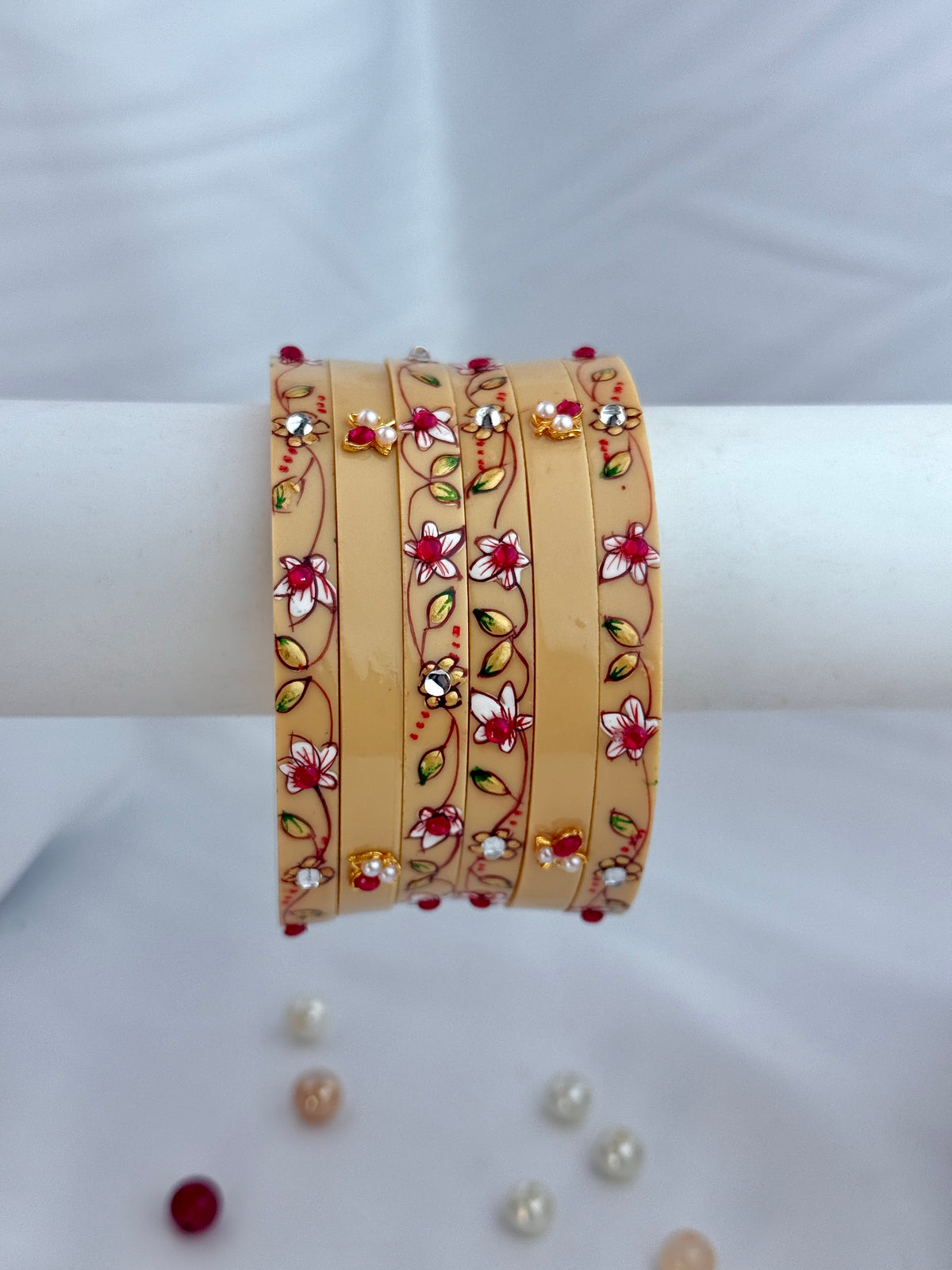 A set of five beige bangles decorated with red and white floral patterns and small gold and red stones, displayed on a white cylindrical stand.