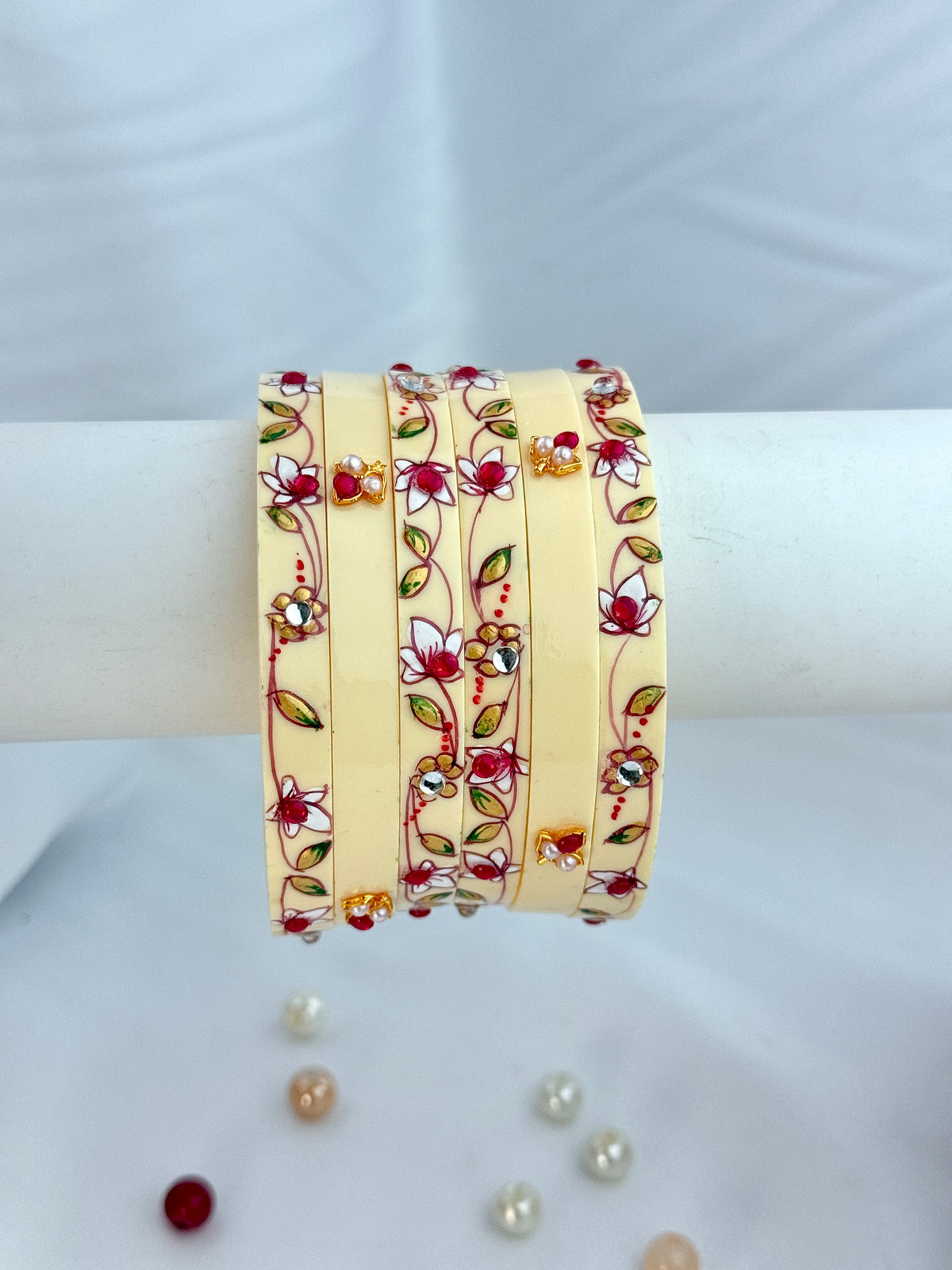 A stack of five cream-colored bangles decorated with red and pink floral patterns, green leaves, and small gold and red stones, displayed on a white stand.