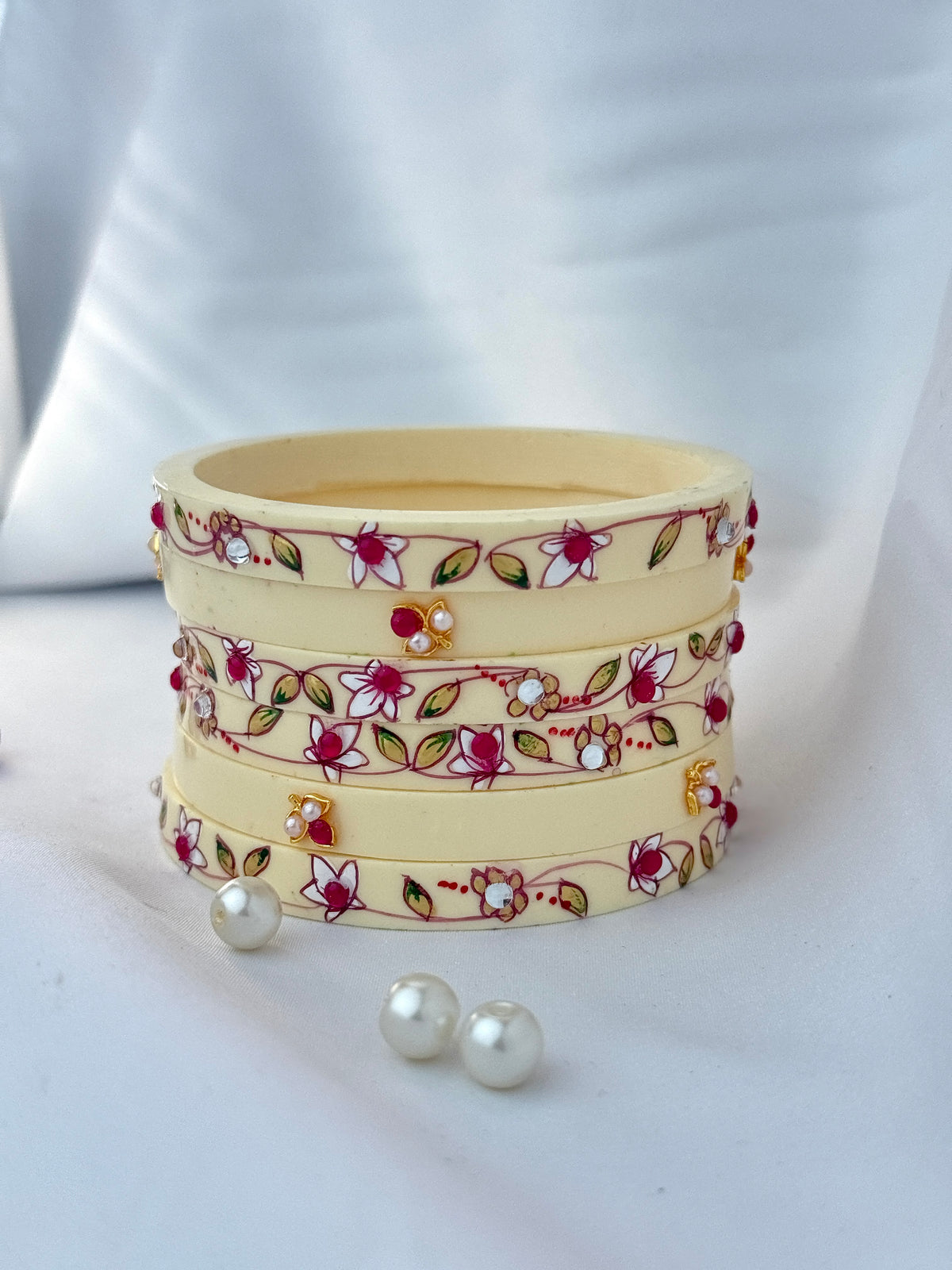 A stack of four cream-colored bangles decorated with red and white floral patterns, green leaves, and small white pearl-like accents, displayed on a white draped fabric background with two loose pearls in front.