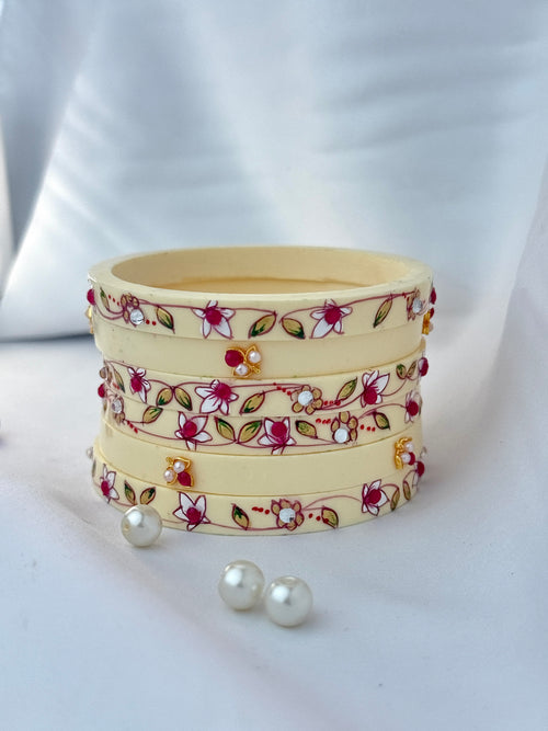 A stack of four cream-colored bangles decorated with red and white floral patterns, green leaves, and small white pearl-like accents, displayed on a white draped fabric background with two loose pearls in front.