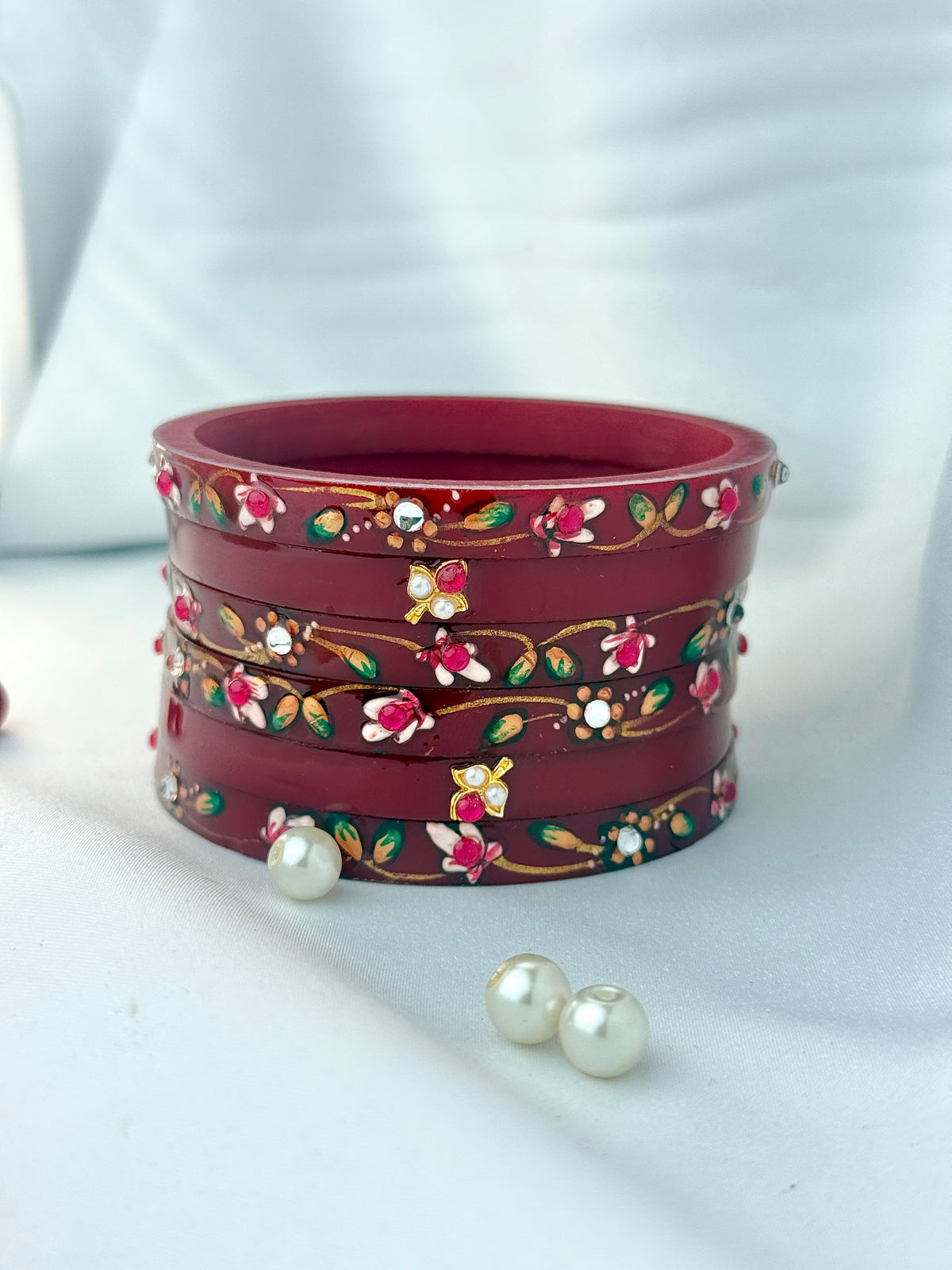 A stack of traditional Indian red bangles decorated with floral and butterfly motifs in gold, pink, green, and white, with two white pearl-like beads placed in front on a white draped fabric background.