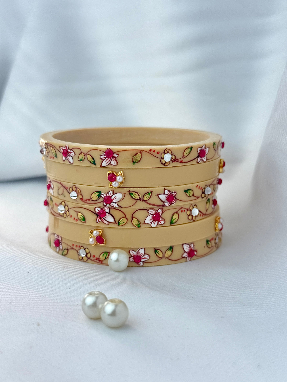 A stack of four tan leather bangles decorated with hand-painted floral motifs in red, pink, green, and white, accented with small gold and white stones. Two loose white pearl earrings are placed in front on a white fabric background.