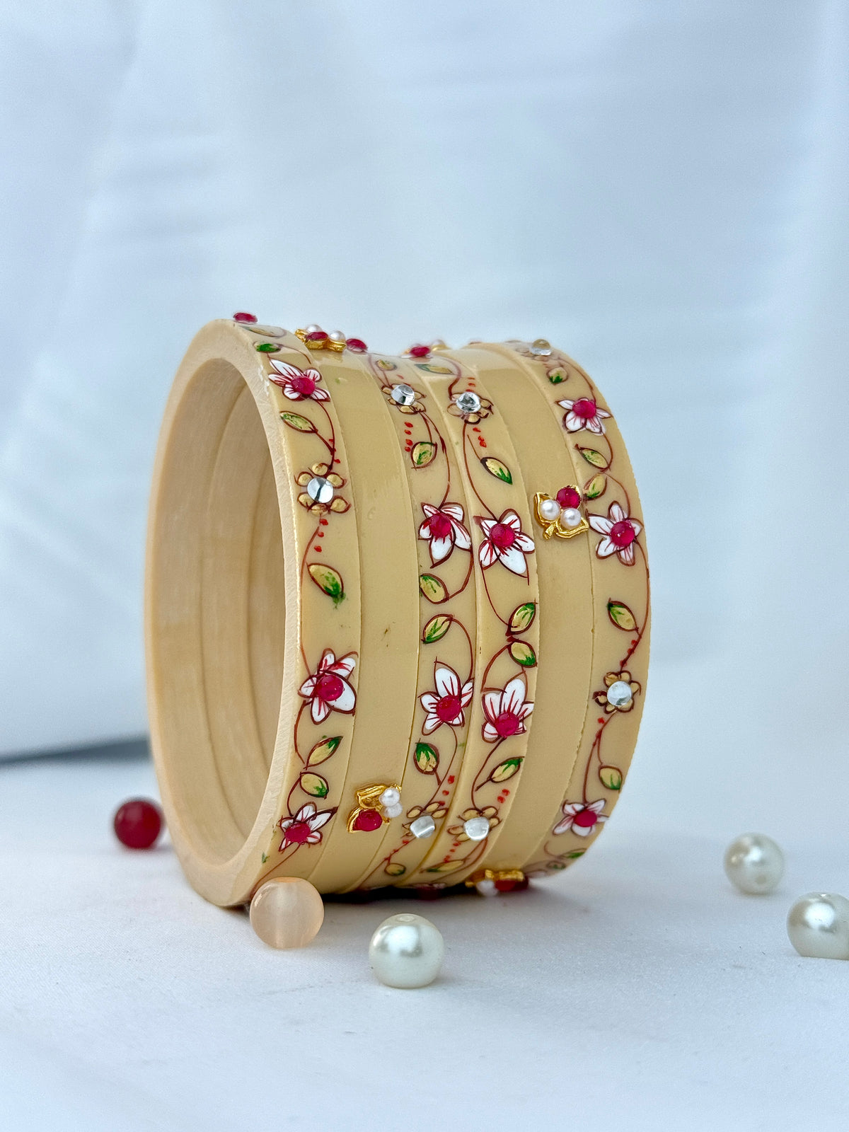 A stack of five light beige wooden bangles decorated with hand-painted floral patterns in red, pink, white, green, and yellow, styled on a white surface with decorative beads.