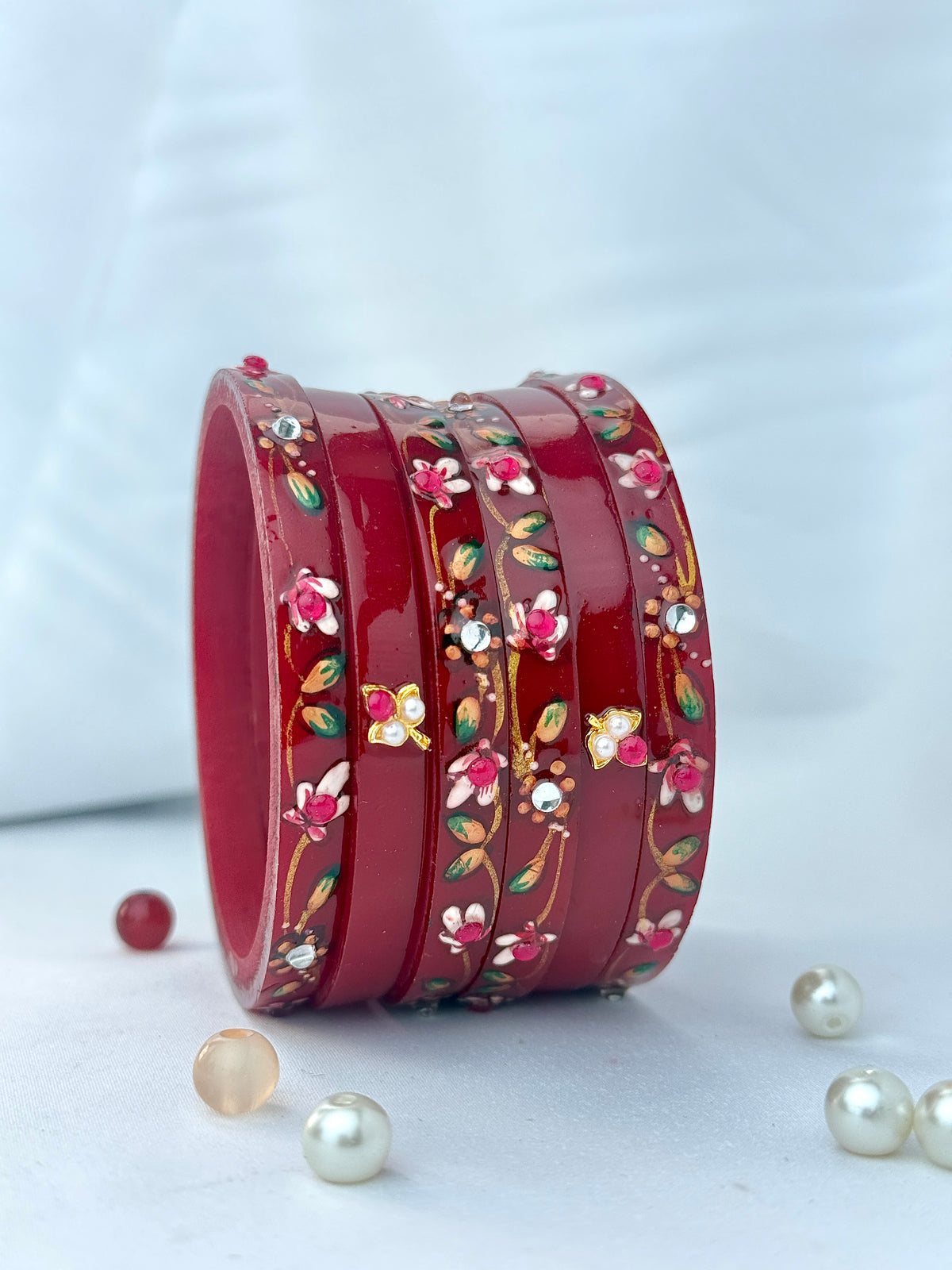 A stack of glossy red bangles decorated with floral and butterfly motifs in pink, green, yellow, and white, arranged on a light surface with scattered pearl-like beads.