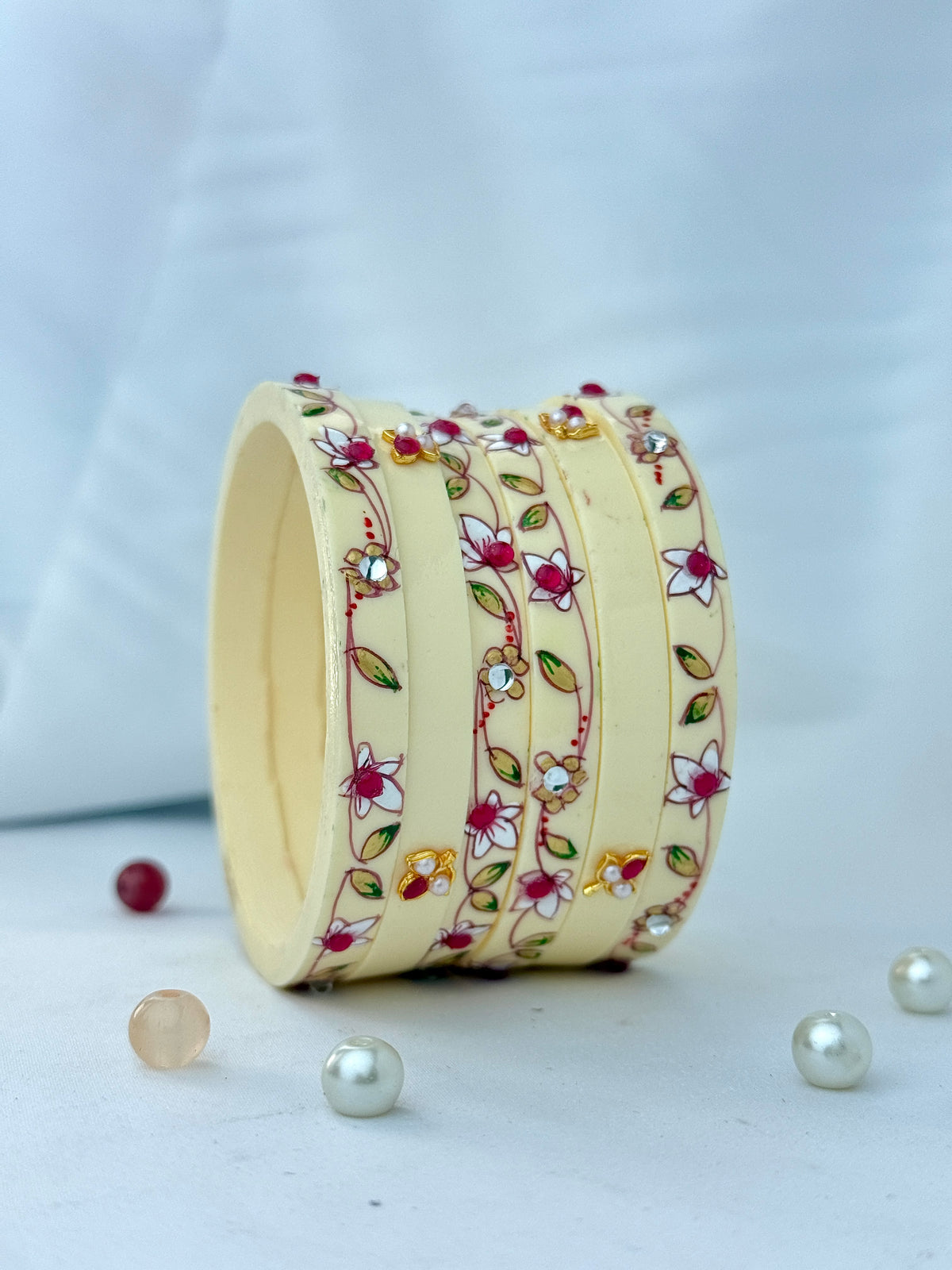 A stack of cream-colored wooden bangles decorated with colorful floral and vine patterns, accented with small rhinestones, displayed on a light surface with decorative pearls and beads.