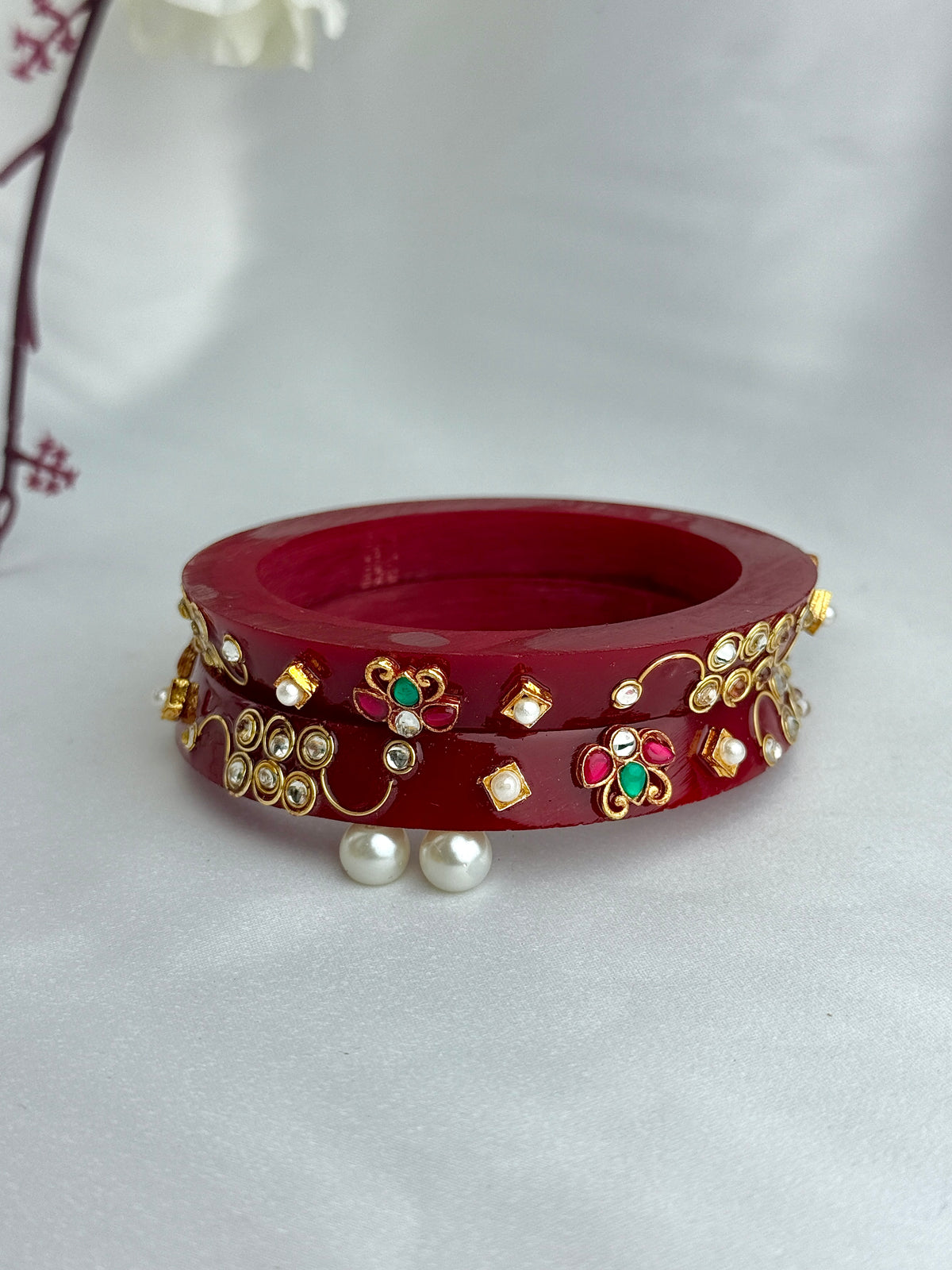 A red bangle bracelet with gold-toned floral and paisley embellishments, decorated with small white stones, green and red enamel details, and two white pearl-like beads hanging in front, set against a white fabric background.