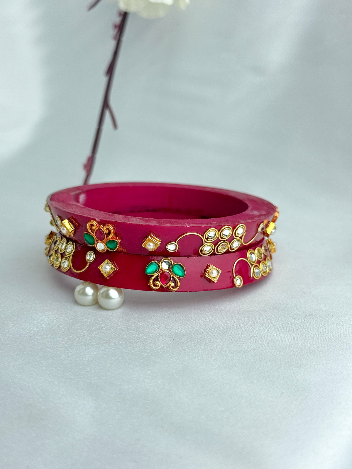 A pair of magenta pink bangles decorated with gold foil, green stones, and white pearl-like accents, displayed on a light gray fabric background.