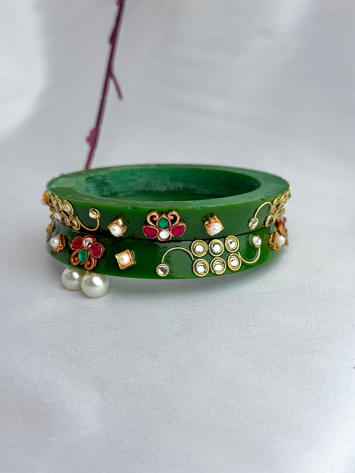 A green bangle bracelet decorated with gold-toned floral and vine motifs, red and green stones, and white faux pearls, displayed on a white fabric background.