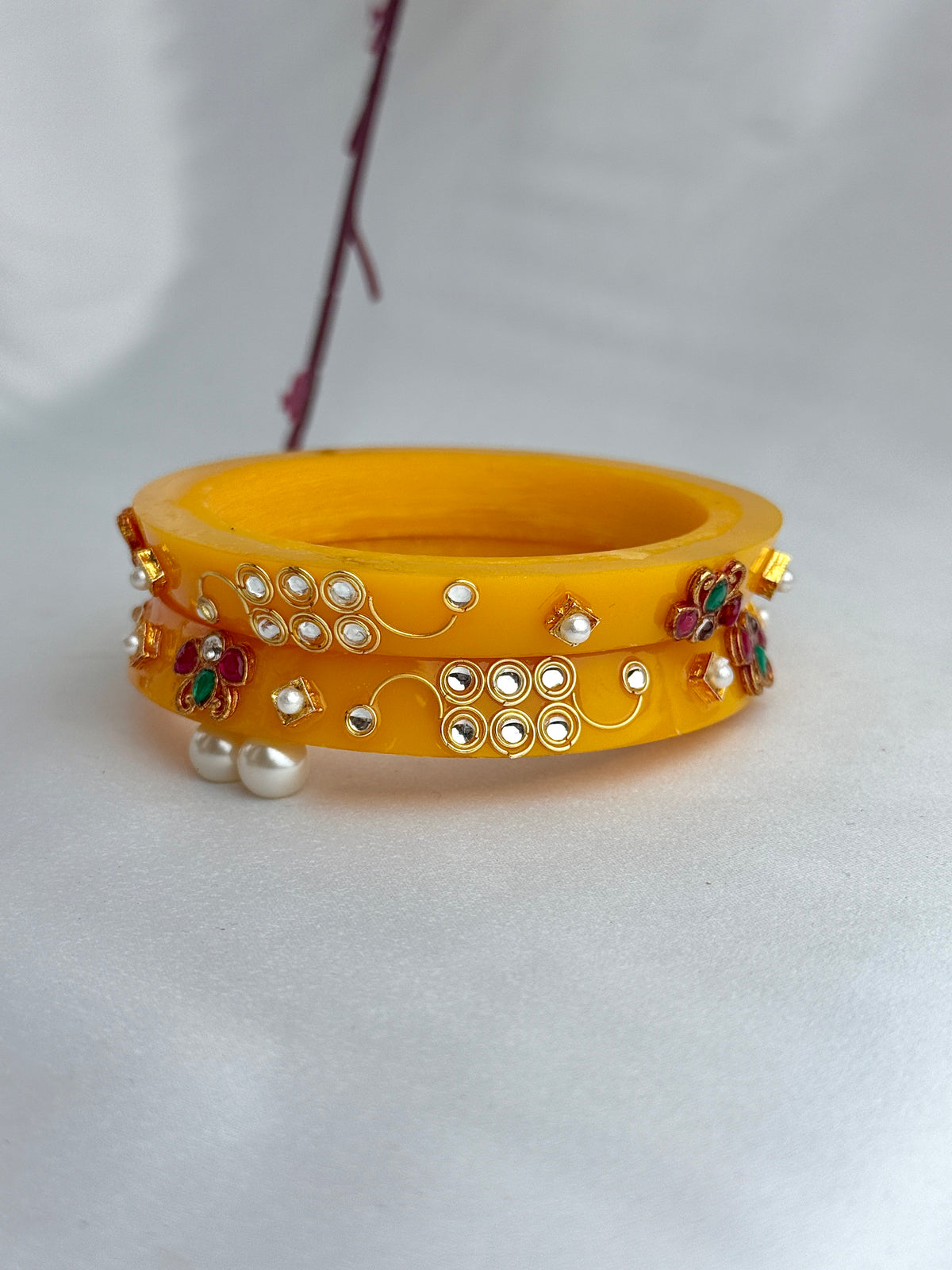 A pair of yellow bangles decorated with gold-toned filigree, small white pearls, and colorful floral embellishments, displayed on a light gray fabric background.
