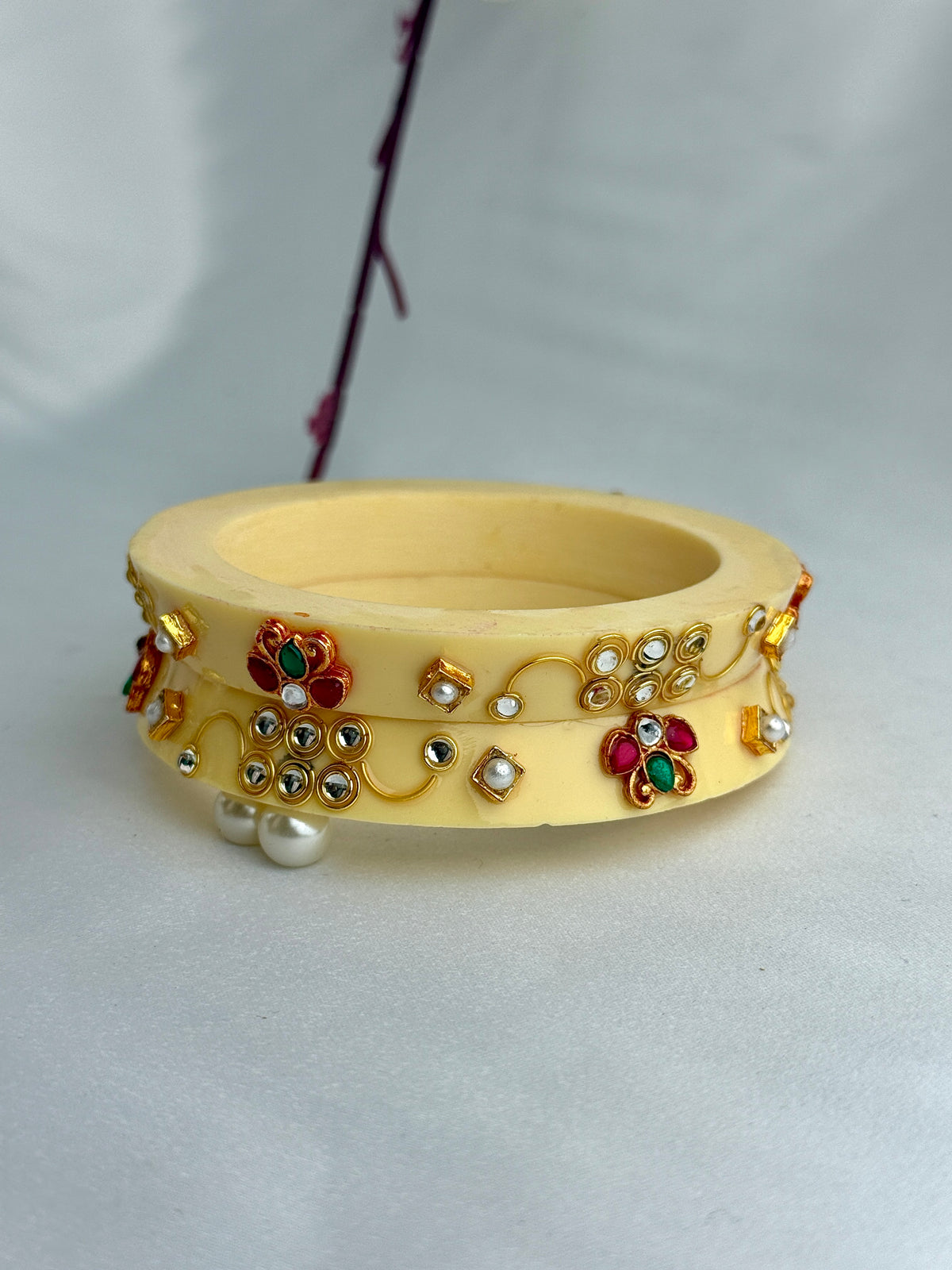 A pale yellow wooden bangle bracelet decorated with gold-toned floral and vine motifs, set with red, green, and white faux gemstones and small pearl-like beads, shown on a light gray background.
