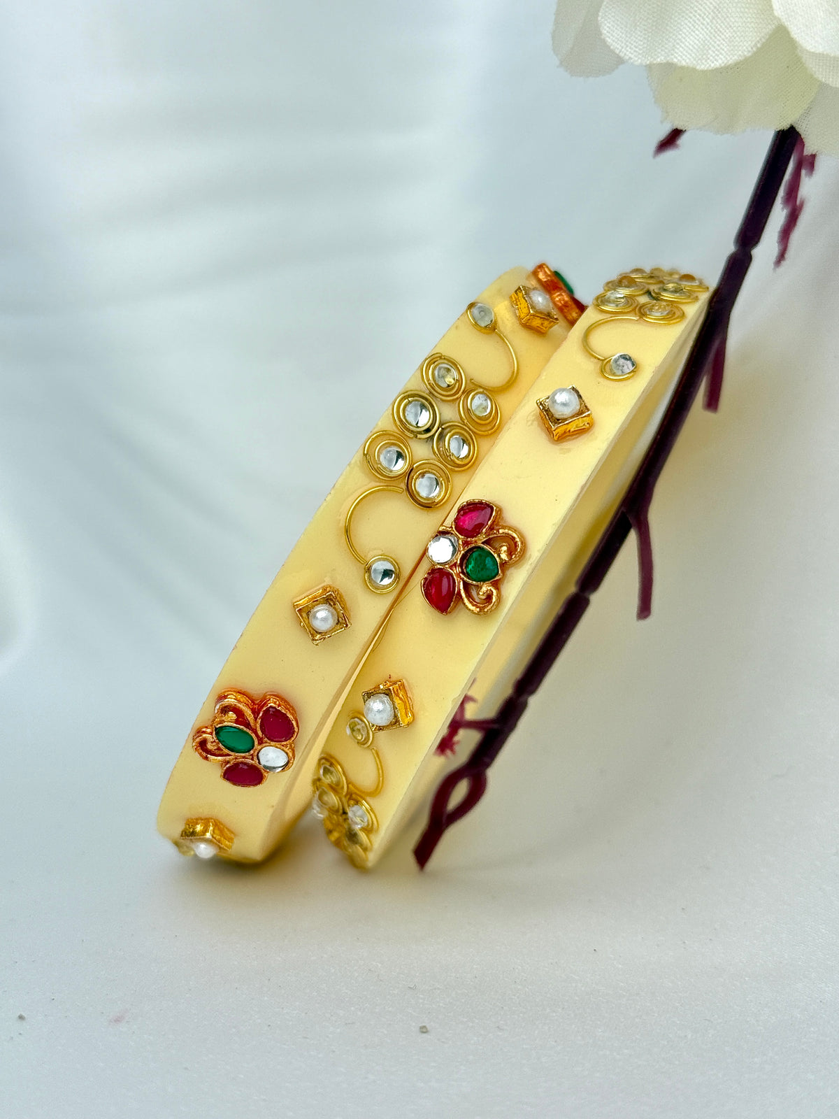 Two gold-toned traditional Indian bangles decorated with small white stones, red and green stones, and gold floral motifs, displayed on a light background with a white flower stem.