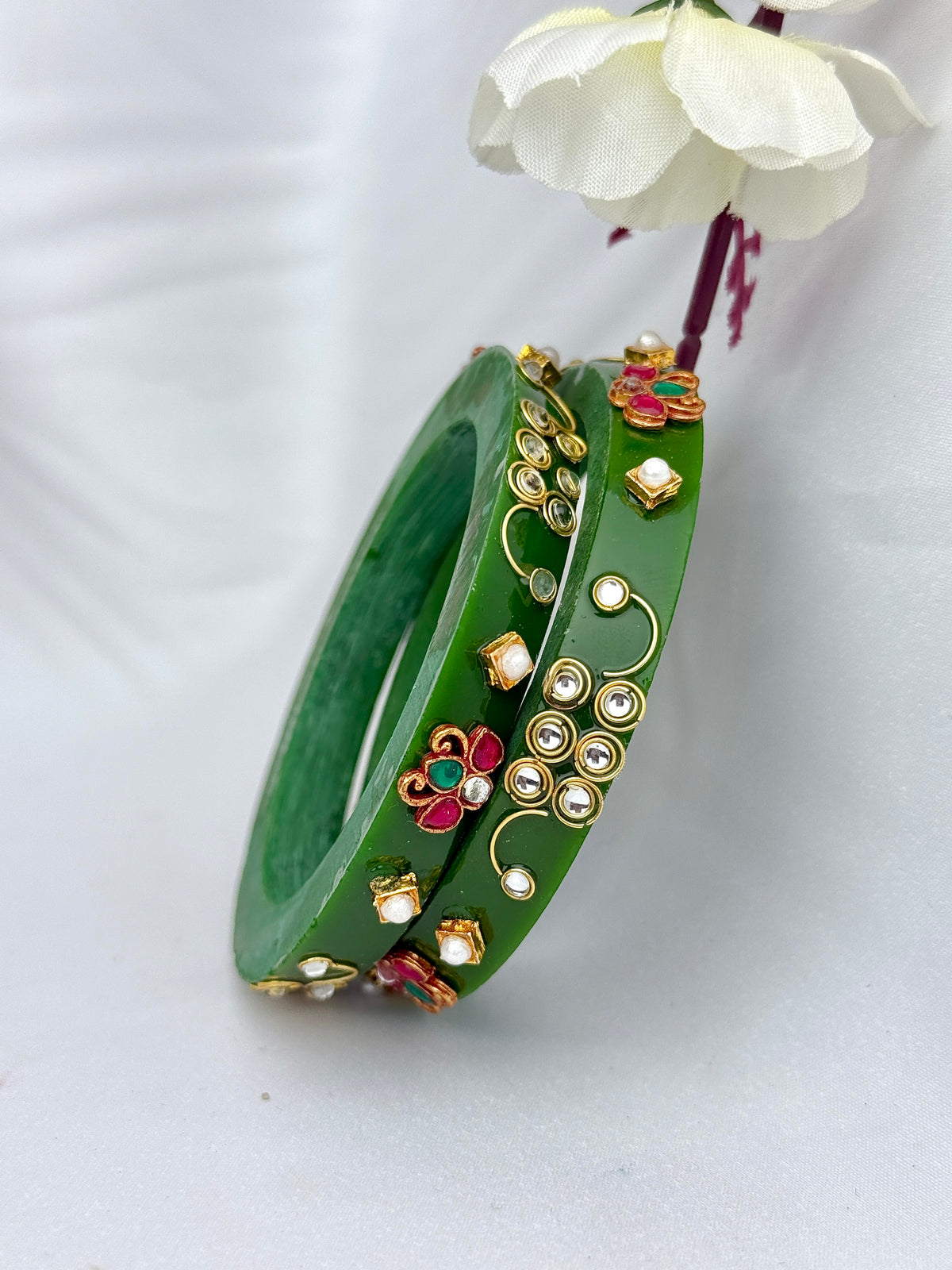 A pair of green bangles decorated with gold-plated metal work, small white stones, and red and green floral accents, displayed on a light gray background with a white flower prop.