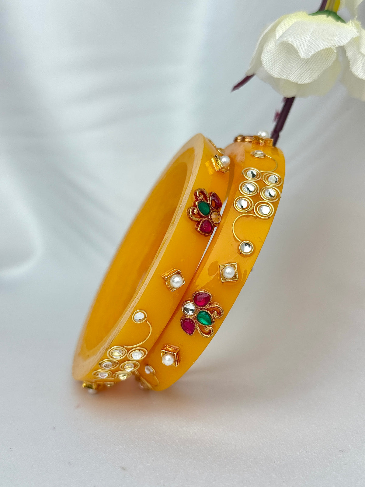 A pair of yellow bangles decorated with gold-toned metalwork, white faux pearls, and small red and green stone floral motifs, photographed on a soft white fabric background with a white flower prop.