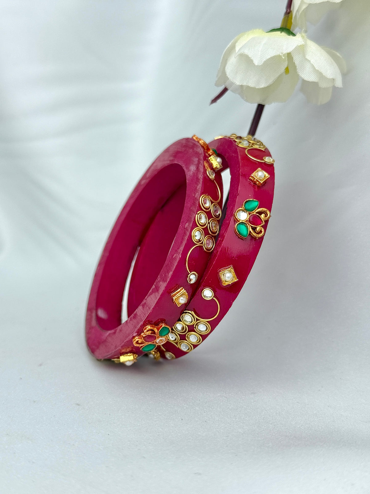 A pair of magenta pink bangles adorned with gold-plated filigree, small white pearl-like studs, and green stone accents, displayed on a soft white background with a white flower prop.