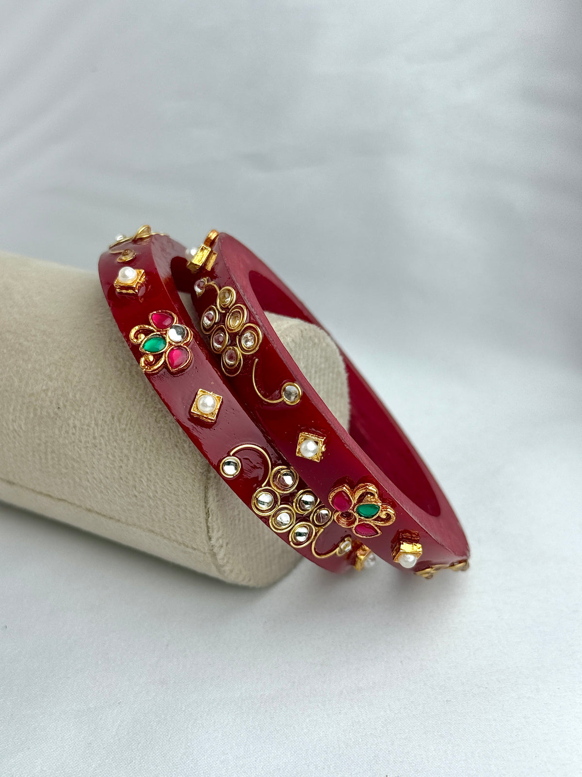 A pair of red bangles decorated with gold-toned floral motifs, small white stones, and green and red enamel details, displayed on a beige jewelry stand against a light gray background.