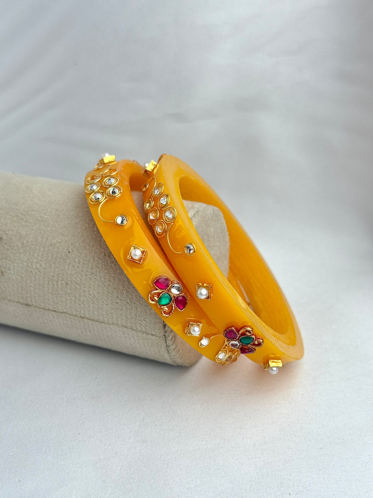 Two bright yellow bangles decorated with small white pearls and colorful floral motifs in red, green, and gold, displayed on a beige jewelry stand against a light gray background.